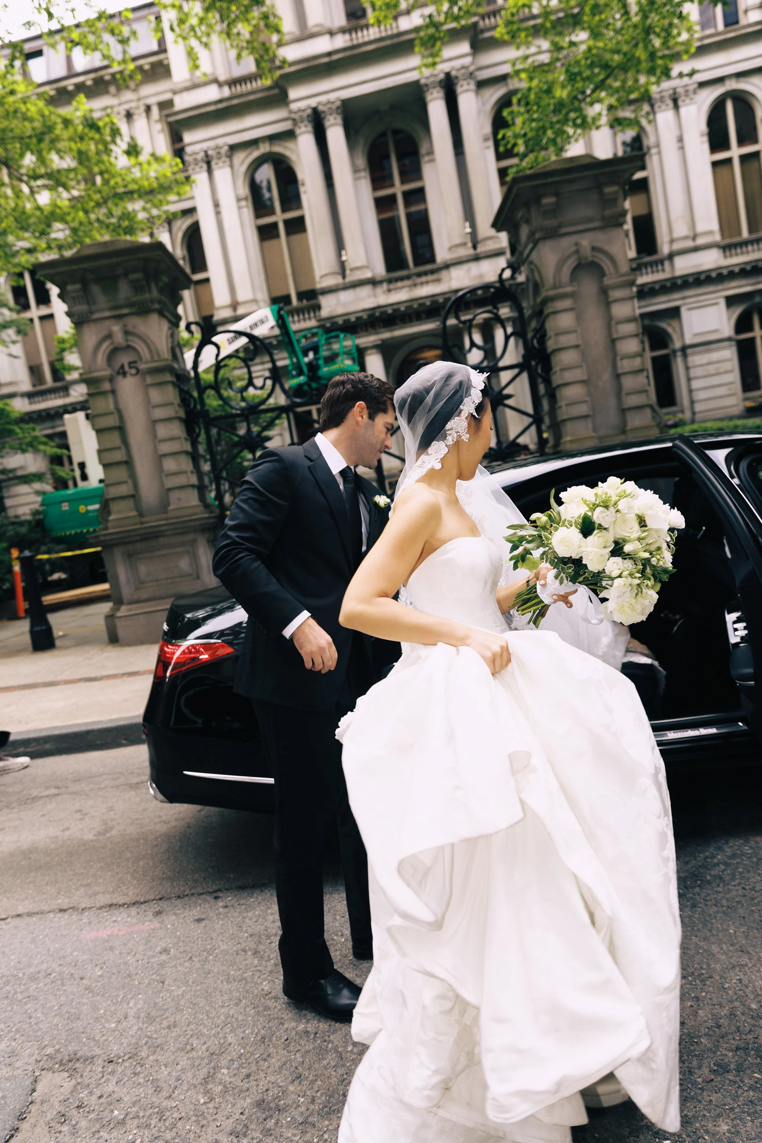 Bride in a white wedding dress and veil holding a bouquet of white flowers, getting into a black car with a man in a dark suit, in front of ornate stone building with green trees.