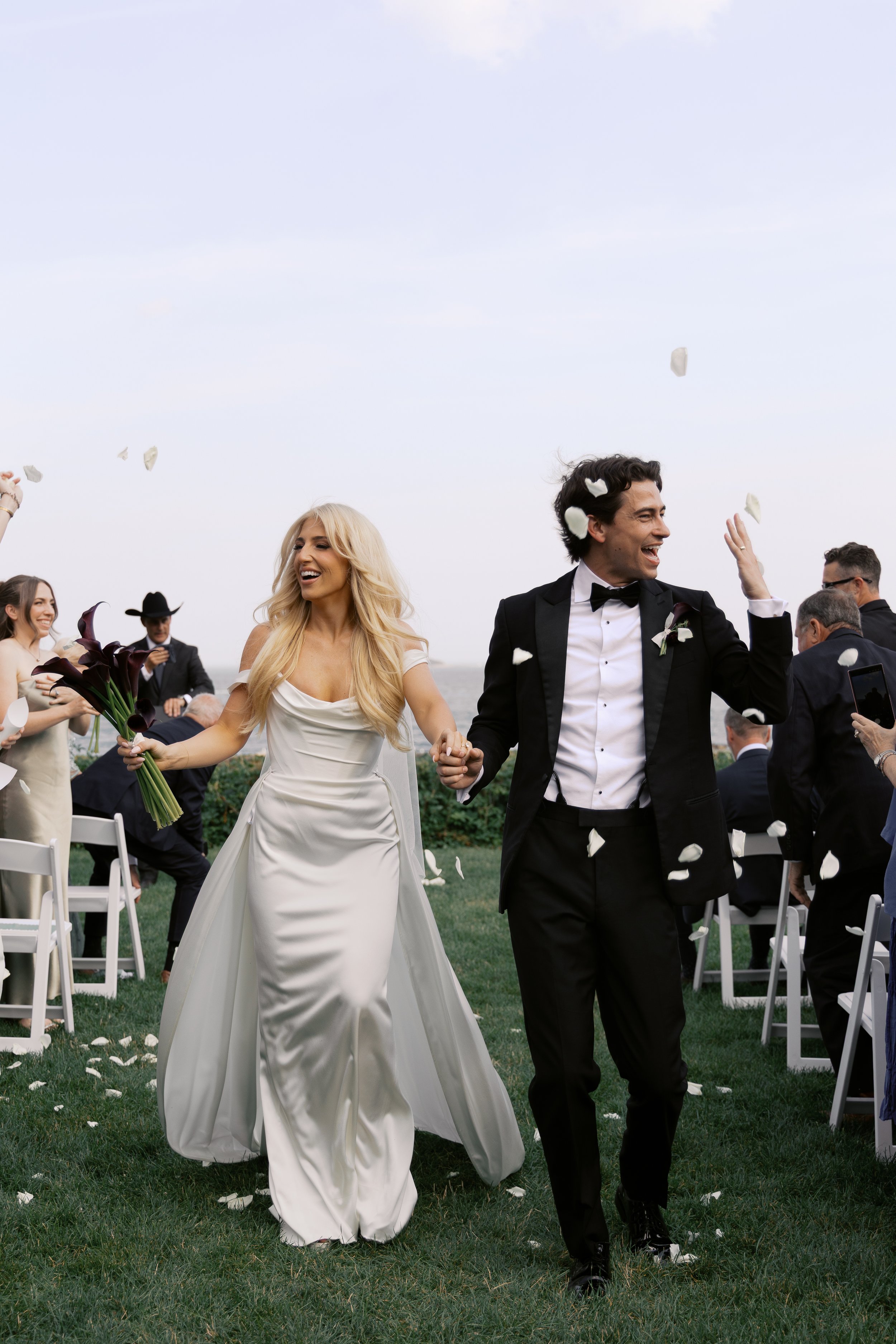 Happy bride and groom walking hand in hand at their wedding reception outdoors, surrounded by guests throwing flower petals.