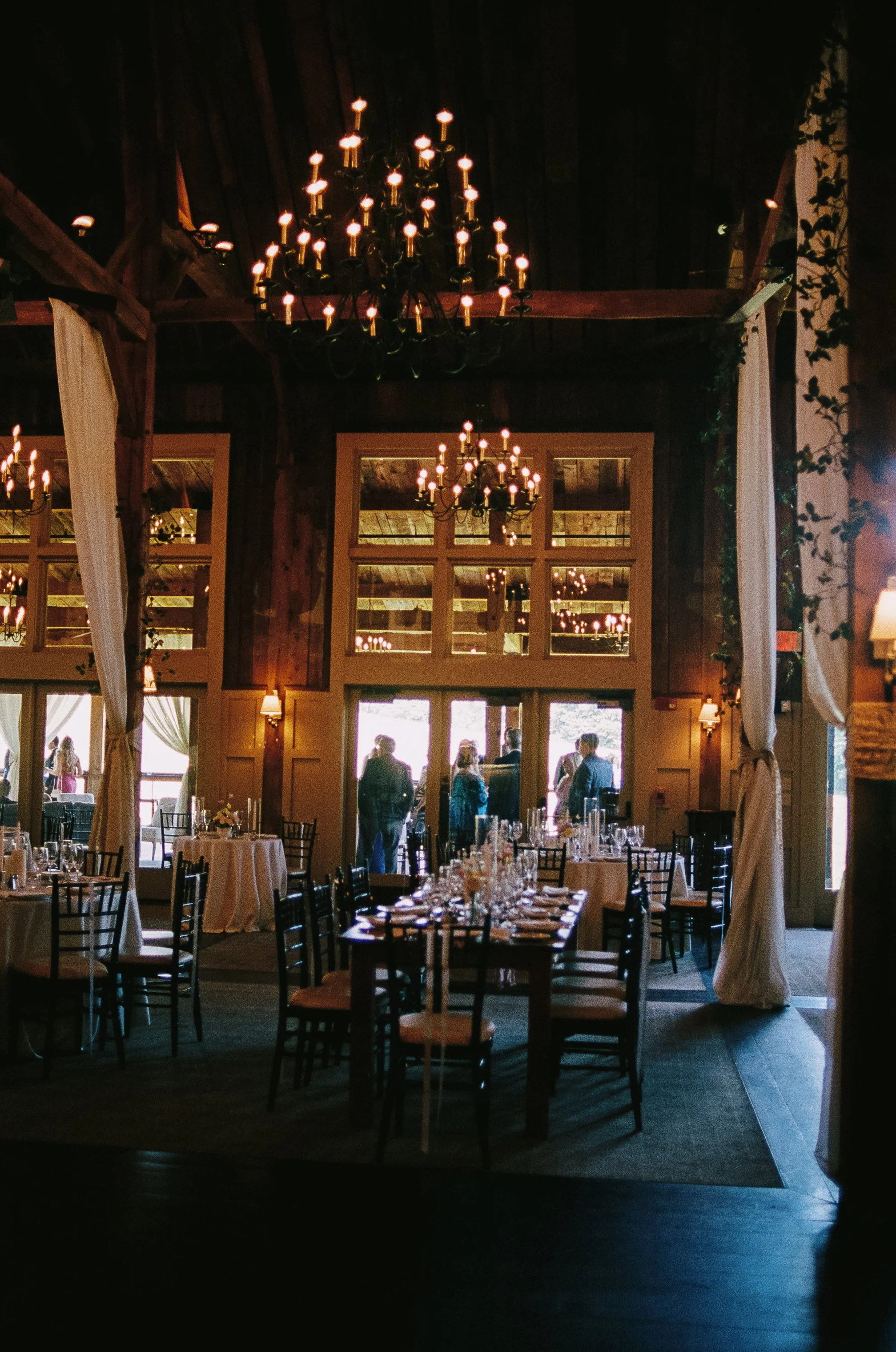 Barn at Gibbet Hill Groton Elegant indoor dining area with wooden walls and ceiling, chandeliers, white curtains, set tables with napkins and glassware, and people standing near the entrance