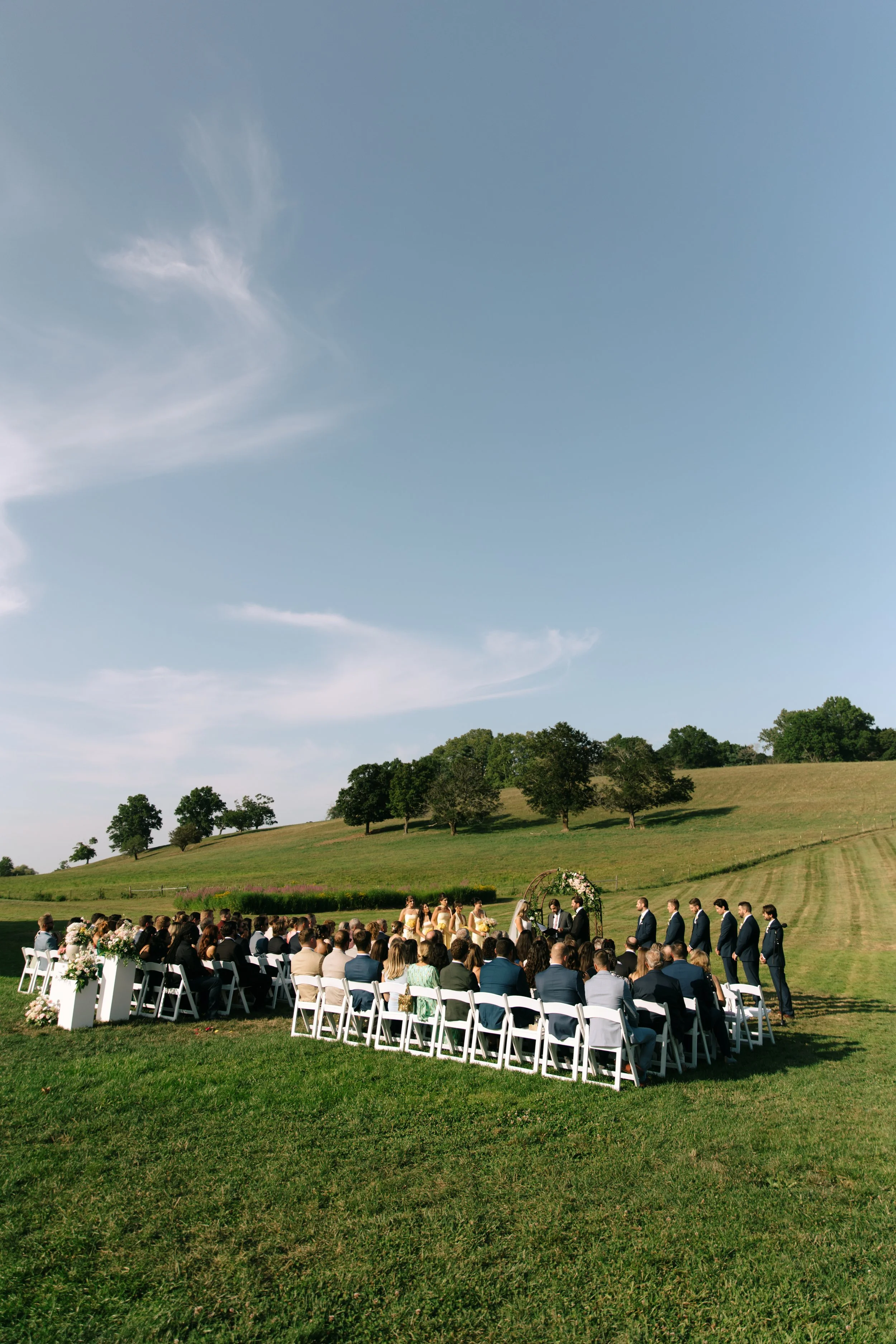 Outdoor wedding ceremony with guests seated on white chairs, a floral arch, and a scenic grassy hillside under a blue sky.