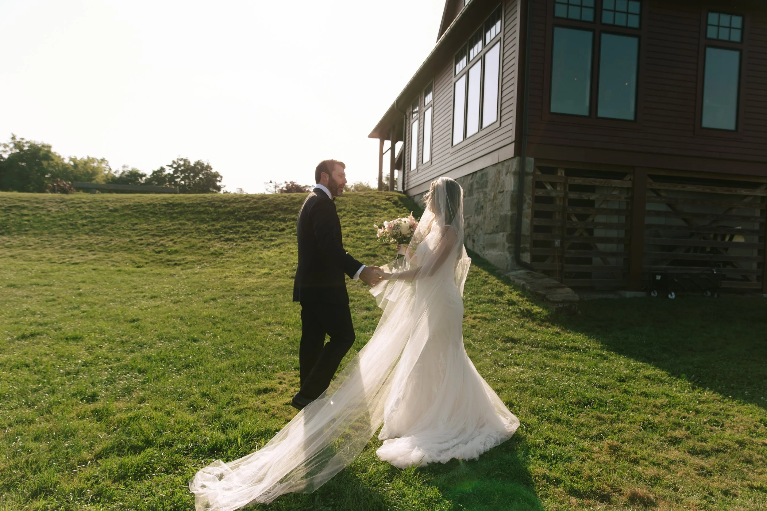 A bride and groom holding hands on a grassy area outside a house during sunset, with the bride wearing a long white gown and veil, and the groom in a black suit, holding a bouquet of flowers.