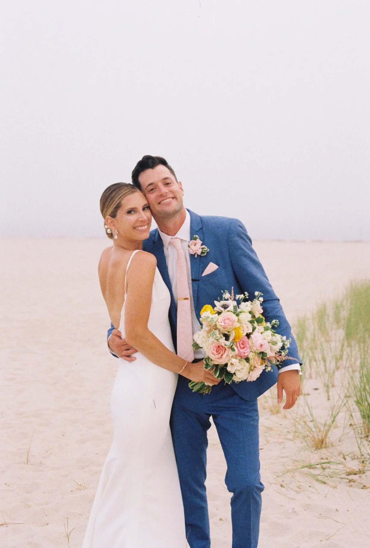 A newlywed couple smiling on a sandy beach, with the bride in a white gown holding a colorful bouquet, and the groom in a blue suit with a pink tie and boutonniere.