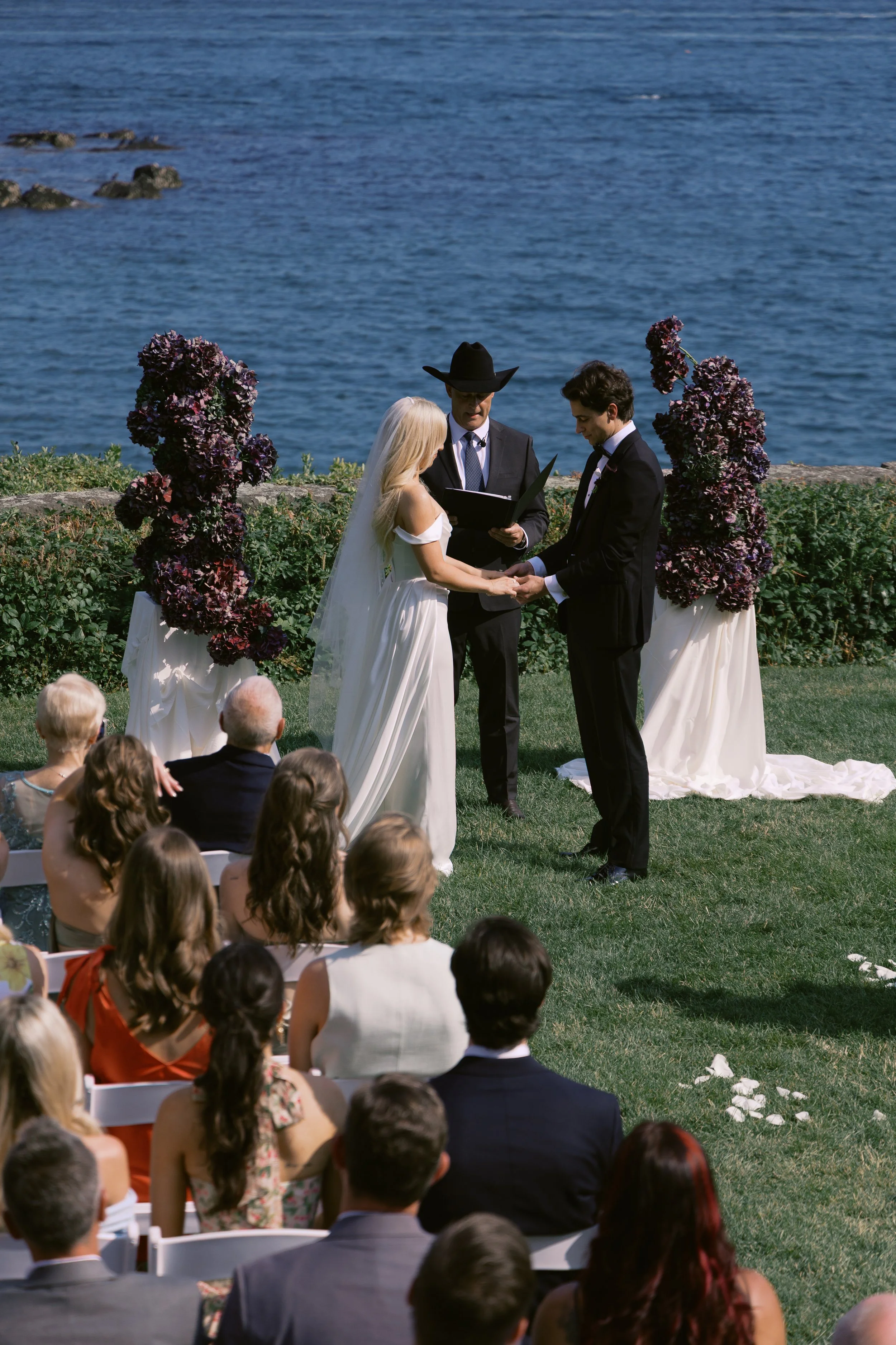 A wedding ceremony taking place outdoors by a body of water with floral decorations and guests seated on grass.