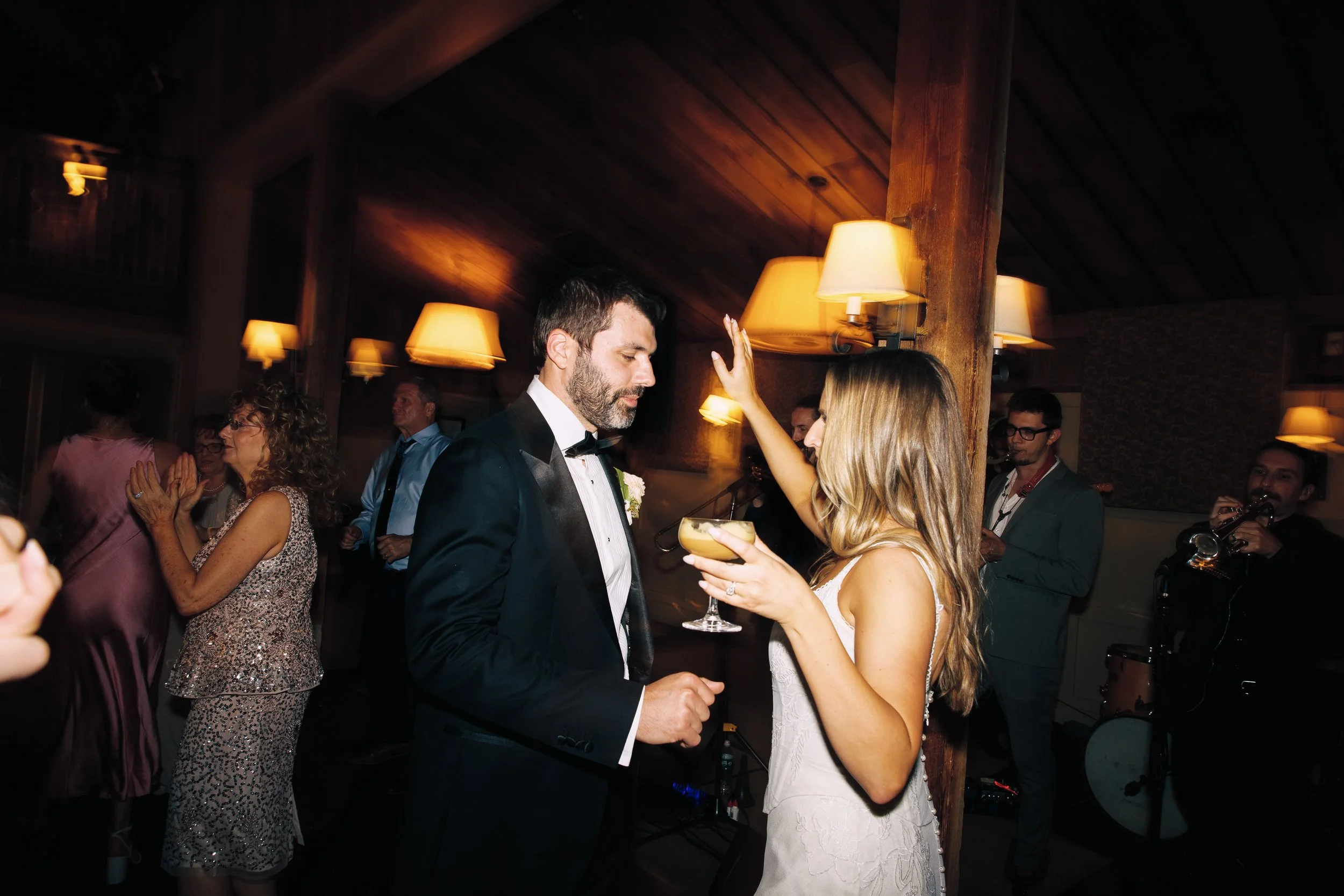 A wedding reception with the bride and groom dancing, surrounded by guests in formal attire, and a live band in the background.