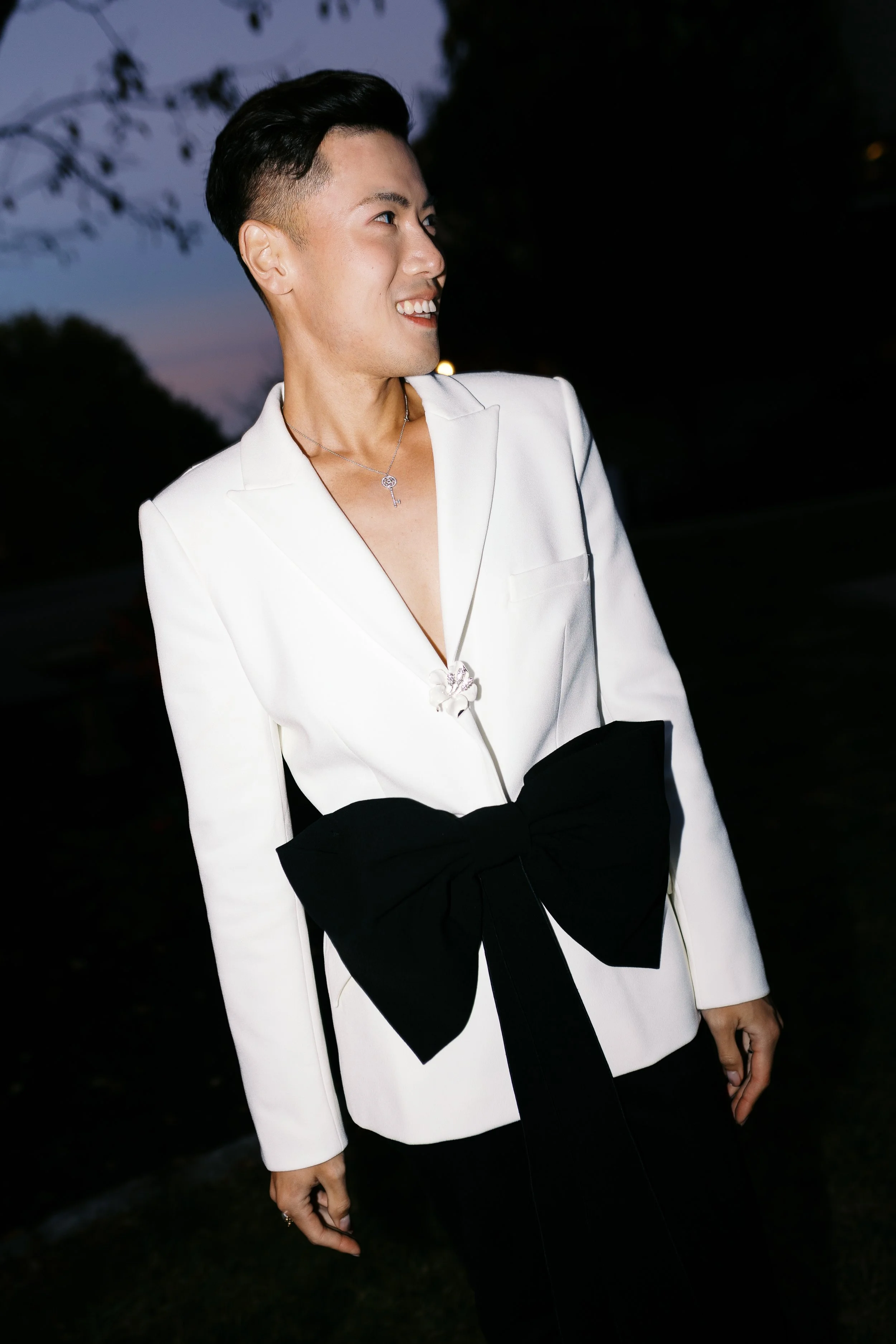 A person wearing a white blazer with a large black bow on the waist, smiling outdoors at dusk.