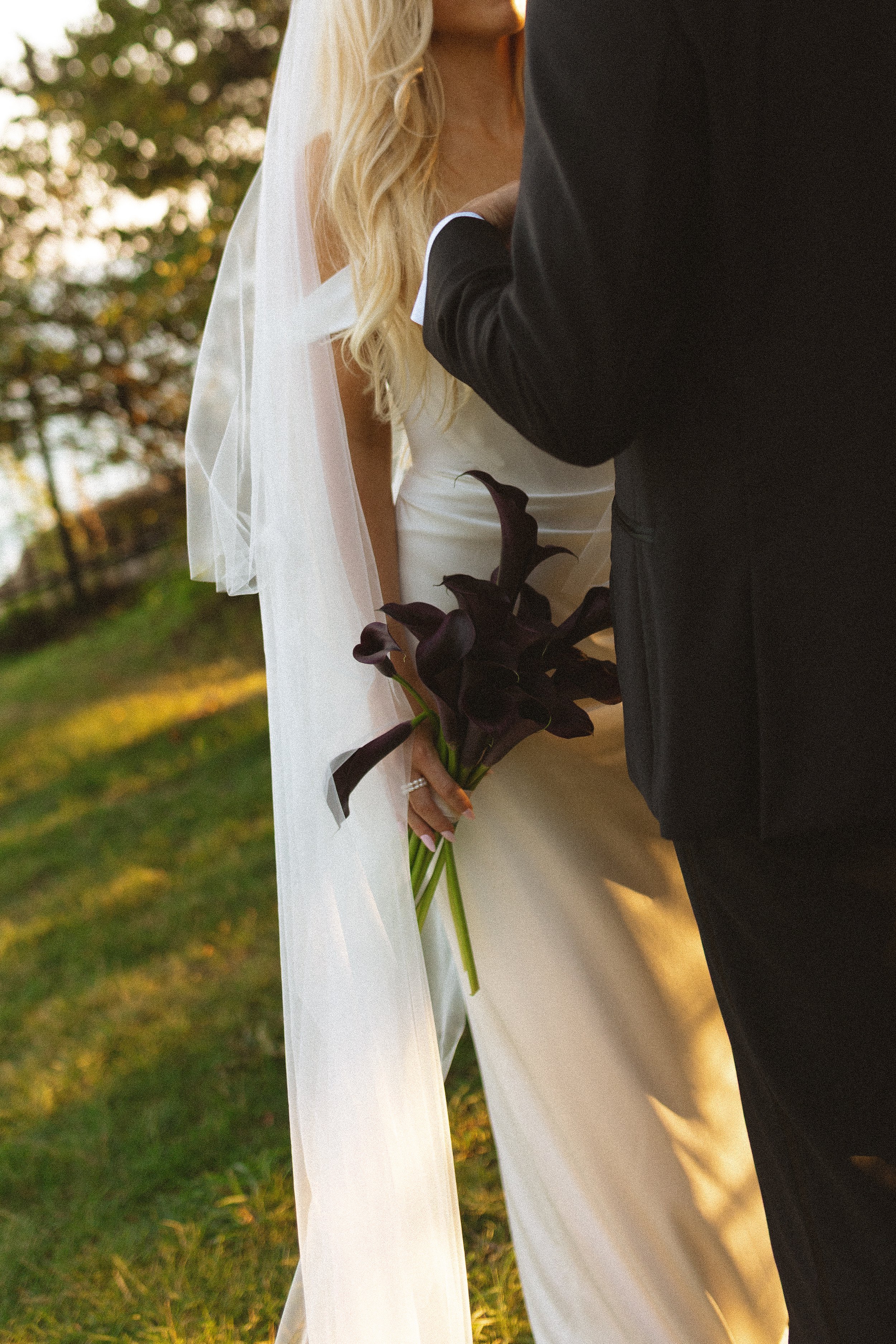 A bride holding a bouquet of dark purple calla lilies standing next to a groom in a black suit at an outdoor wedding ceremony.