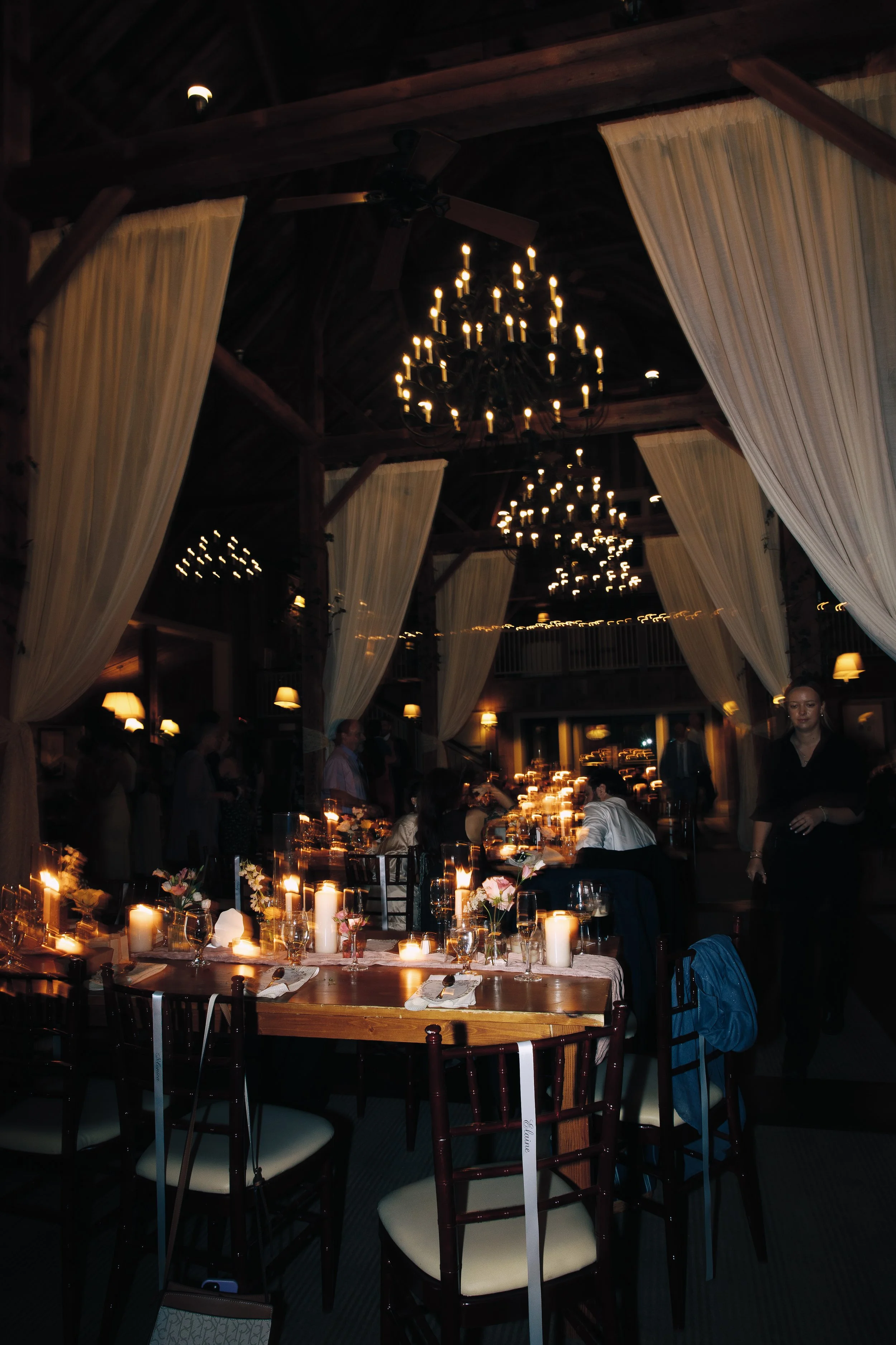 Dimly lit banquet hall with long table set for dinner, decorated with flowers, candles, and glassware. Chandeliers hang from dark wooden ceiling. Cream-colored curtains hang from the ceiling, and there are people standing and sitting in the backgroun