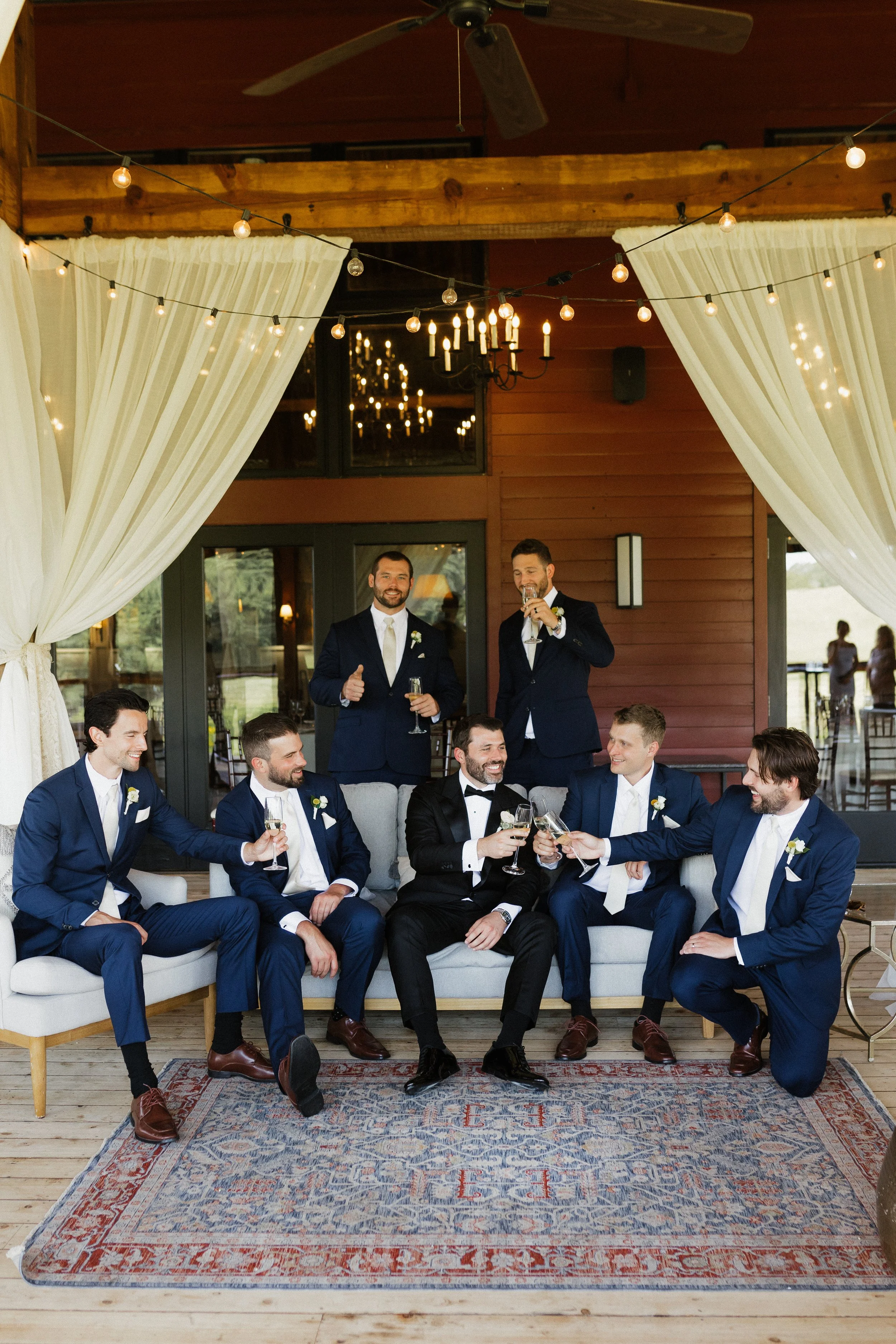 A groom and six groomsmen celebrating at a wedding reception, sitting and standing on a sofa with drinks, in a rustic venue decorated with string lights and curtains.