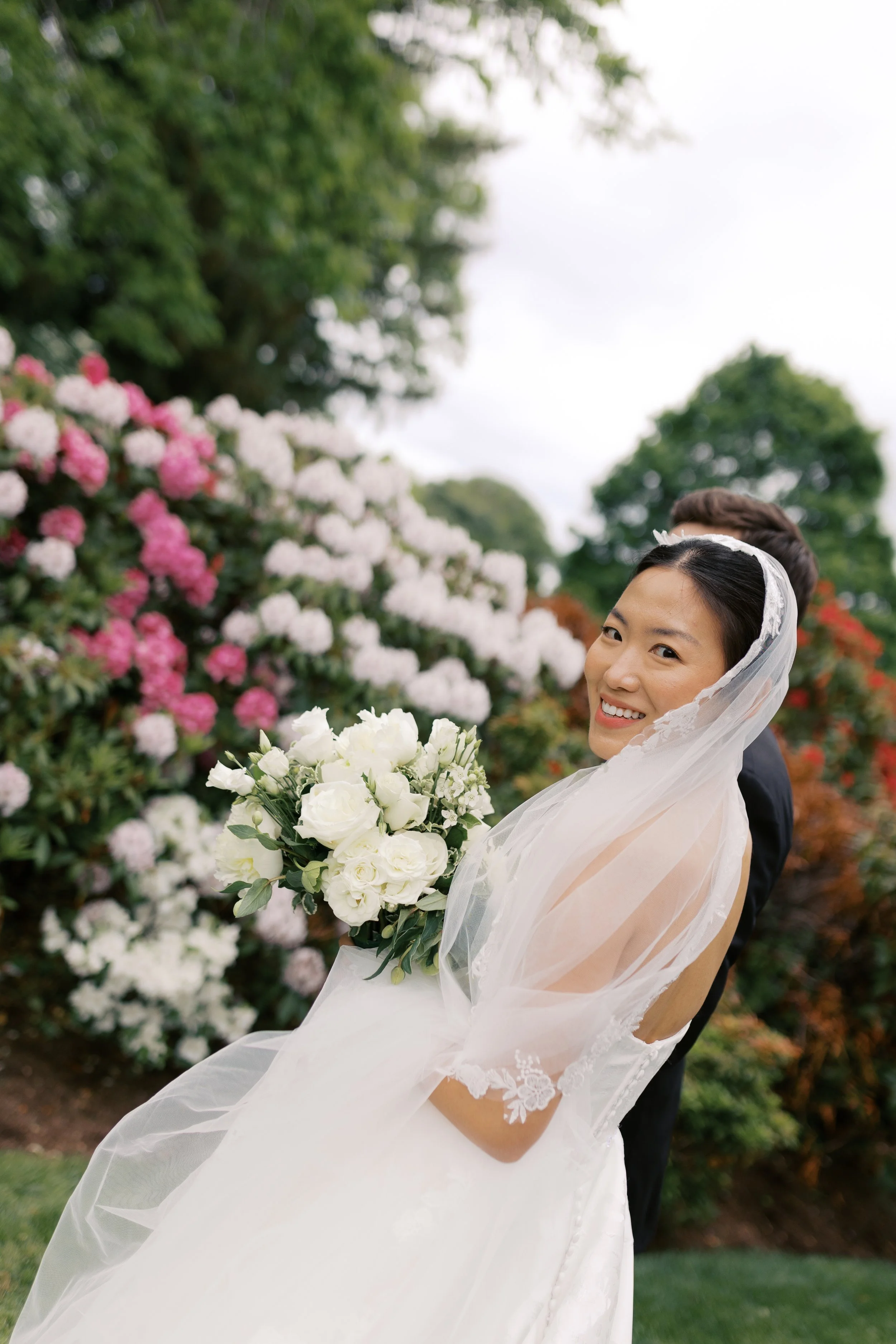 Bride in a wedding dress holding a bouquet of white roses, smiling, outdoors with pink and white flowers and green trees in the background.
