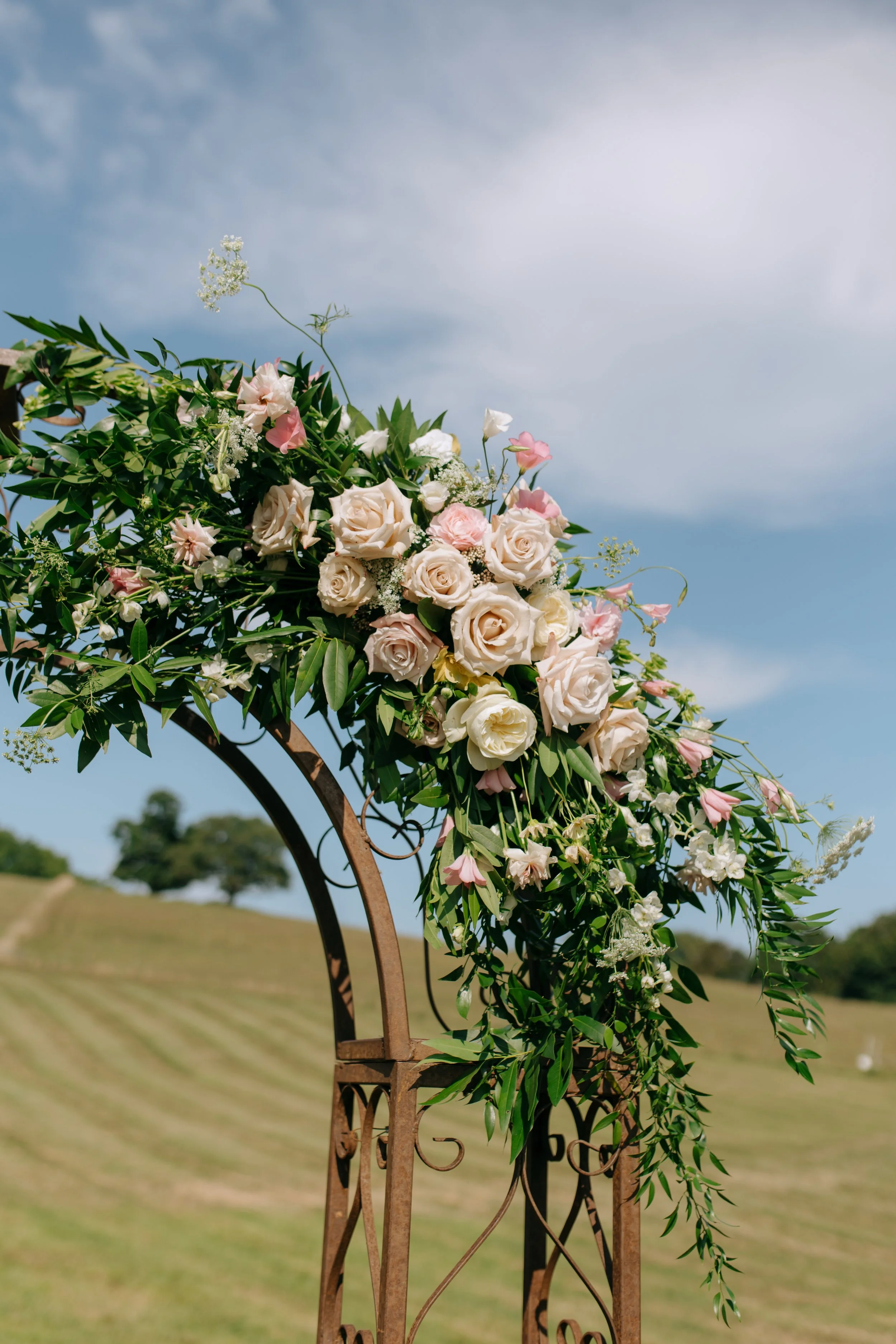 Flowers arranged on a metal arch at an outdoor wedding ceremony with a grassy field and trees in the background under a cloudy sky.