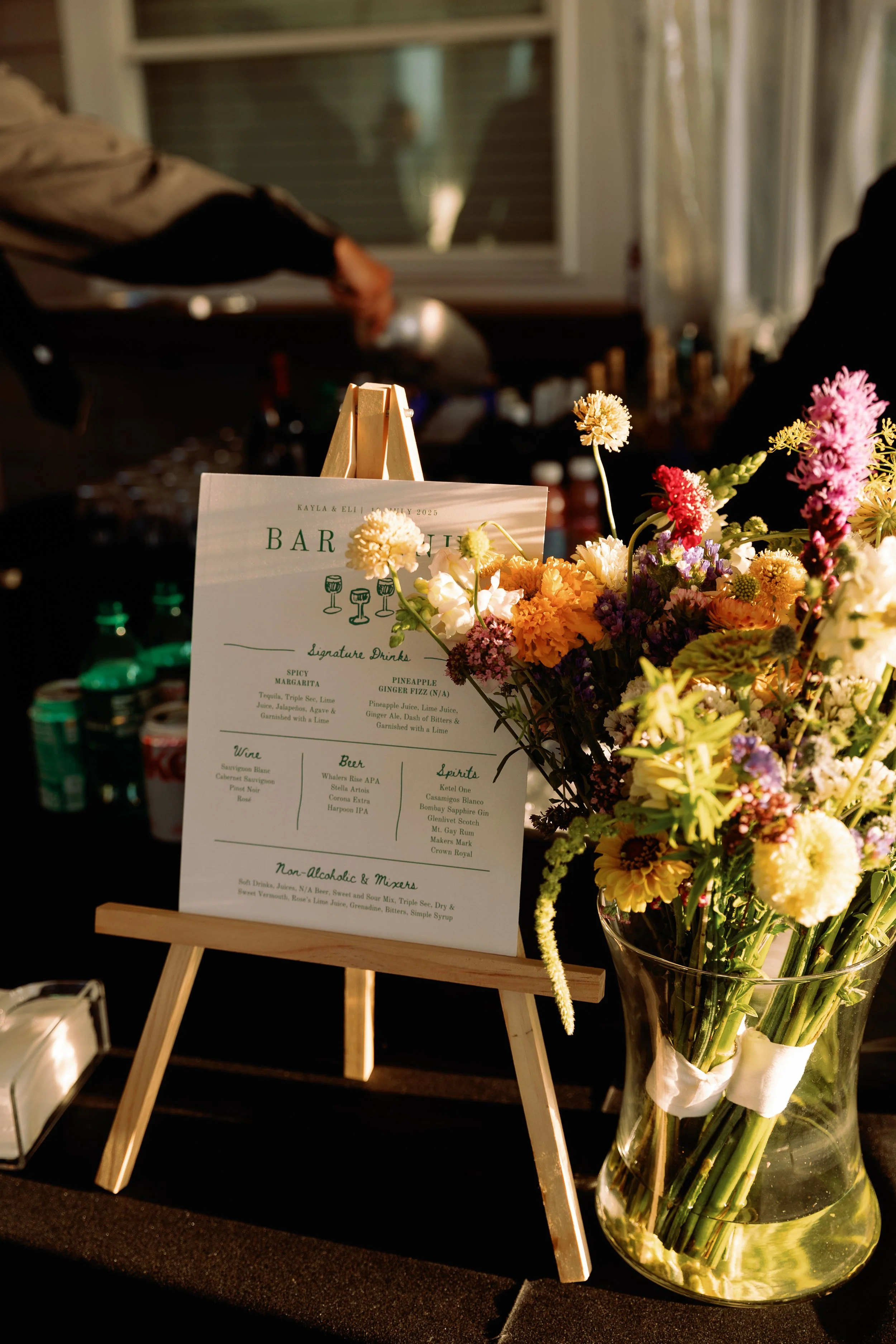 A drink menu on a small wooden easel next to a large bouquet of colorful flowers in a glass vase, at a bar or event setting