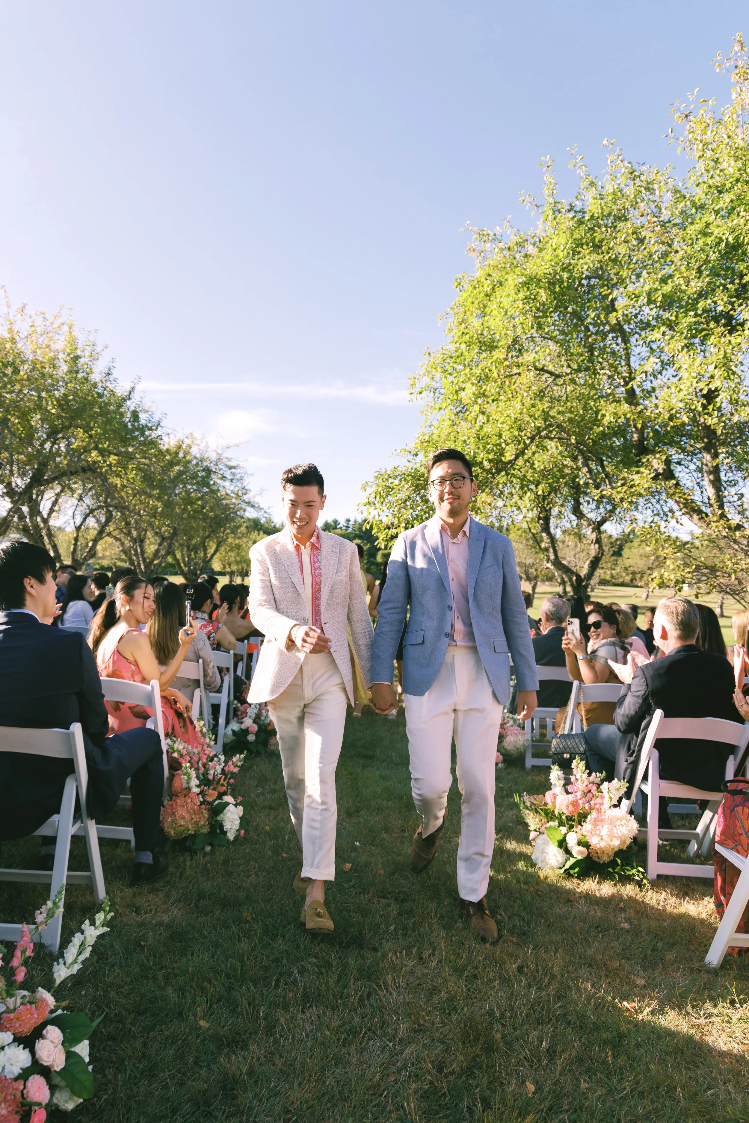 Gay couple holding hands and walking down an aisle at an outdoor wedding ceremony, with guests seated on either side, under a clear blue sky and surrounded by green trees.