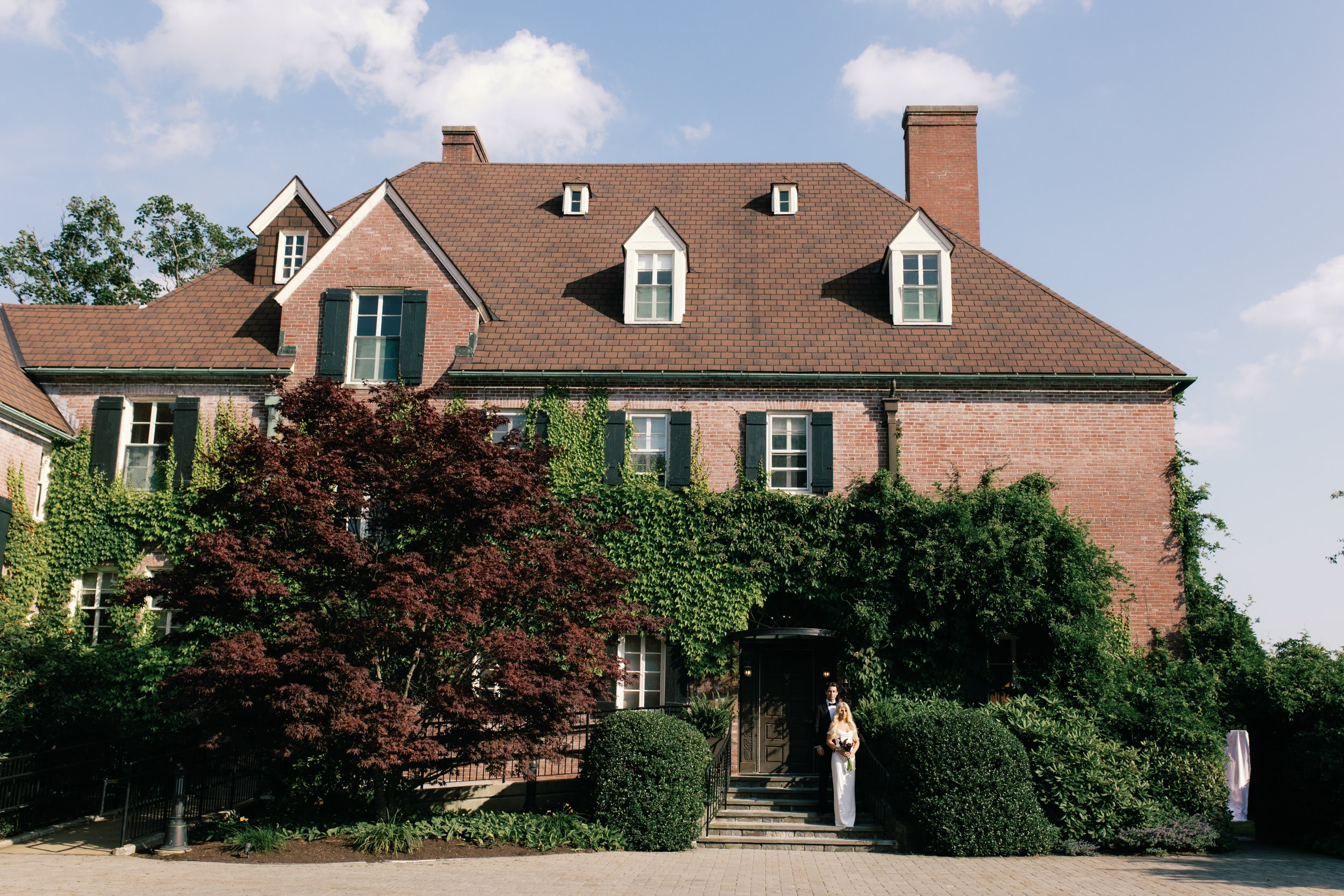A large brick house with a red tile roof, surrounded by green trees and bushes. Two people are standing at the front door, dressed formally, possibly for a wedding.