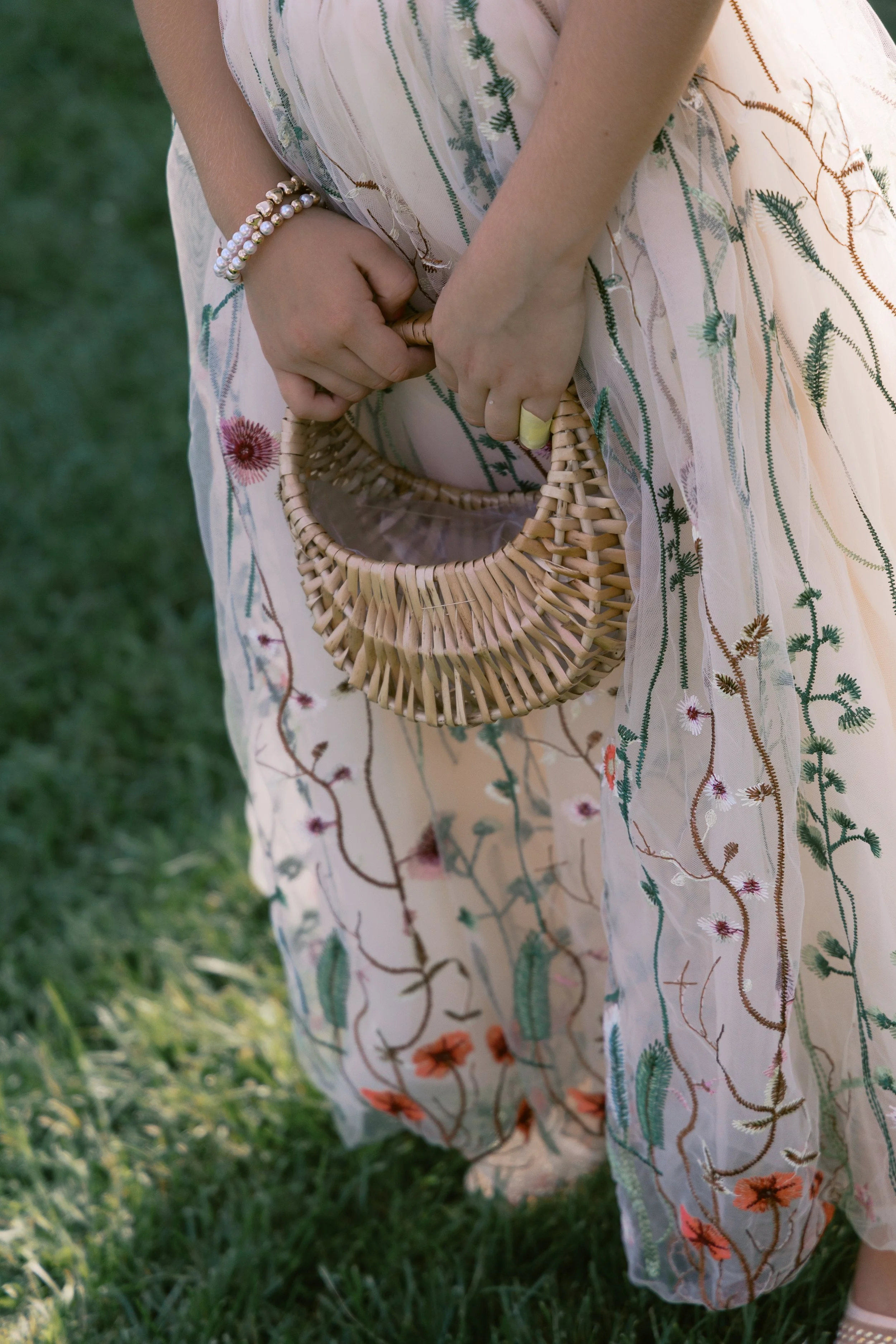 Close-up of a woman wearing a floral embroidered dress, holding a small wicker handbag, standing on grass.