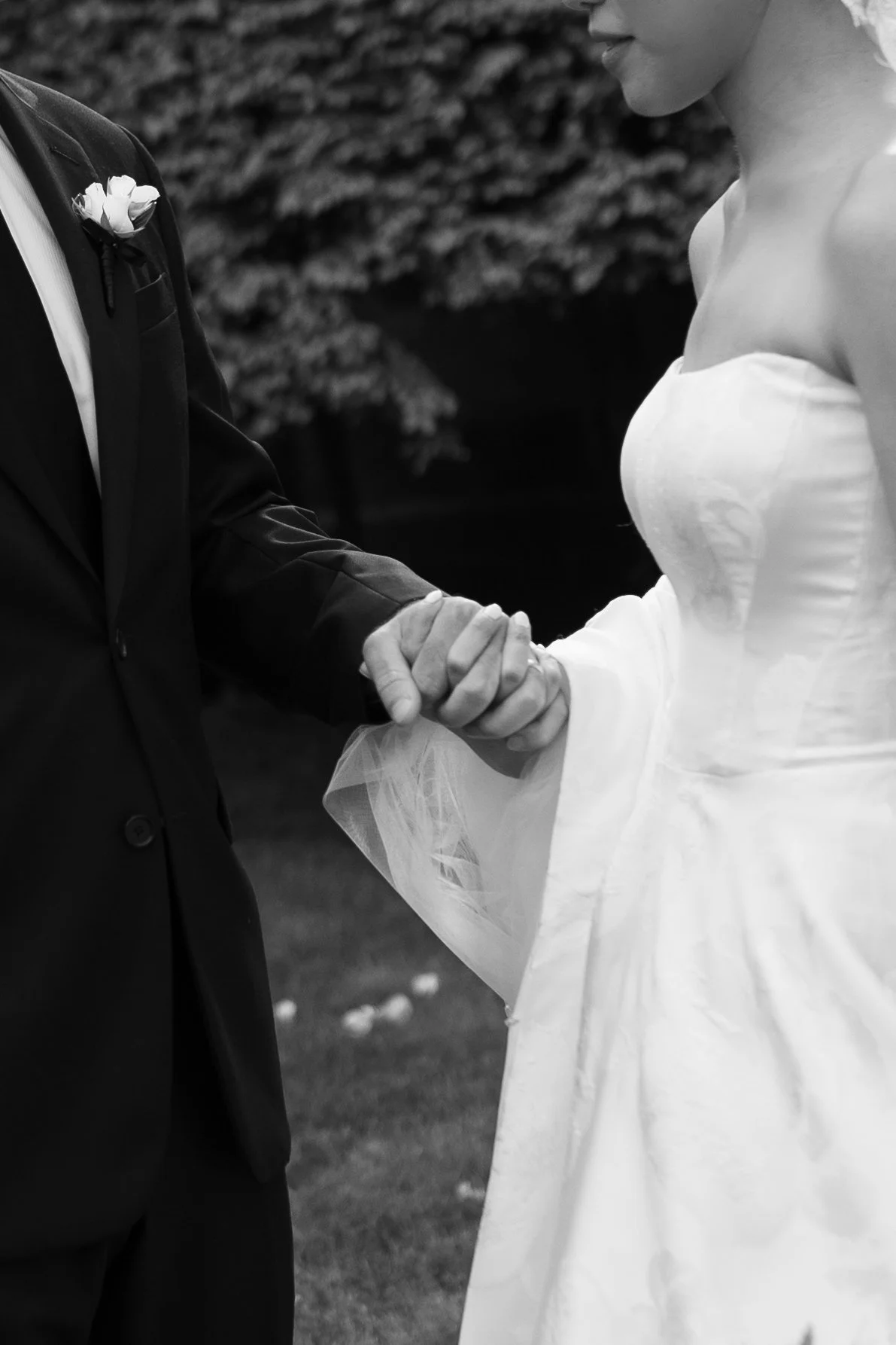 A black-and-white photo of a bride and groom holding hands during a wedding ceremony outdoors.