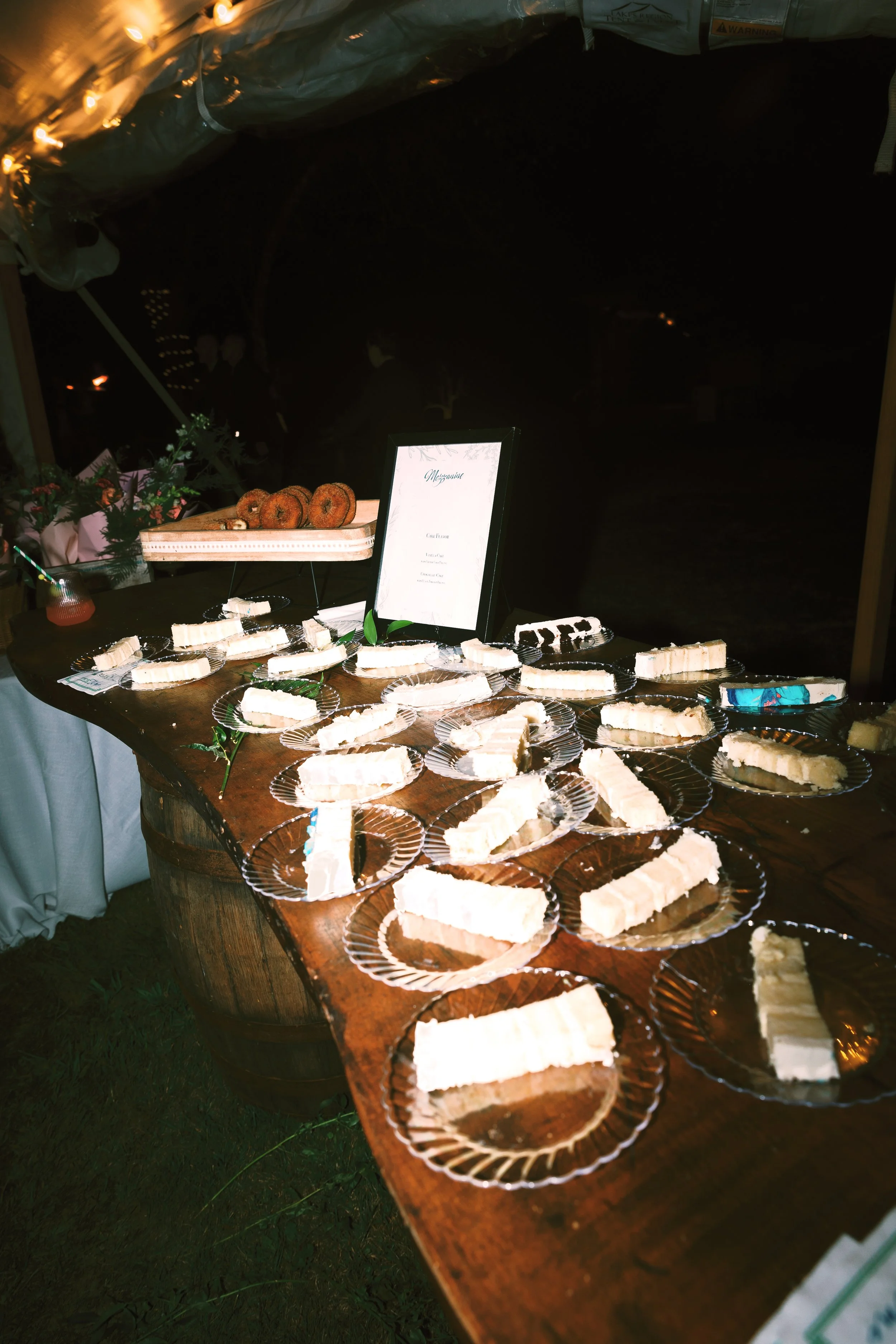 Cheesecake slices arranged on glass plates on a wooden table at a wedding reception, with a framed menu, floral decorations, and a plate of donuts in the background.