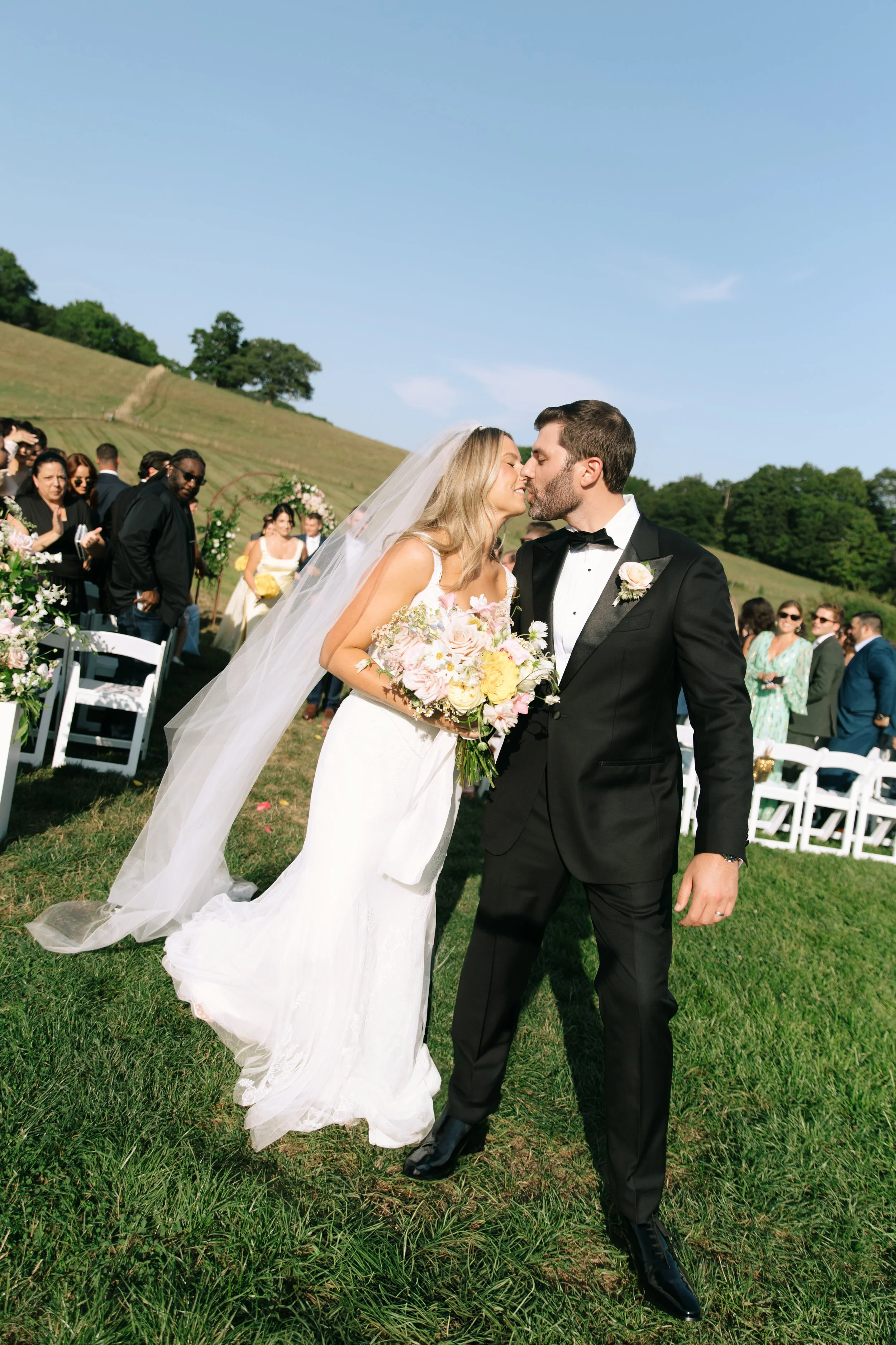 A bride and groom kiss during their outdoor wedding ceremony on a grassy hill under a clear blue sky, with guests seated and standing in the background.