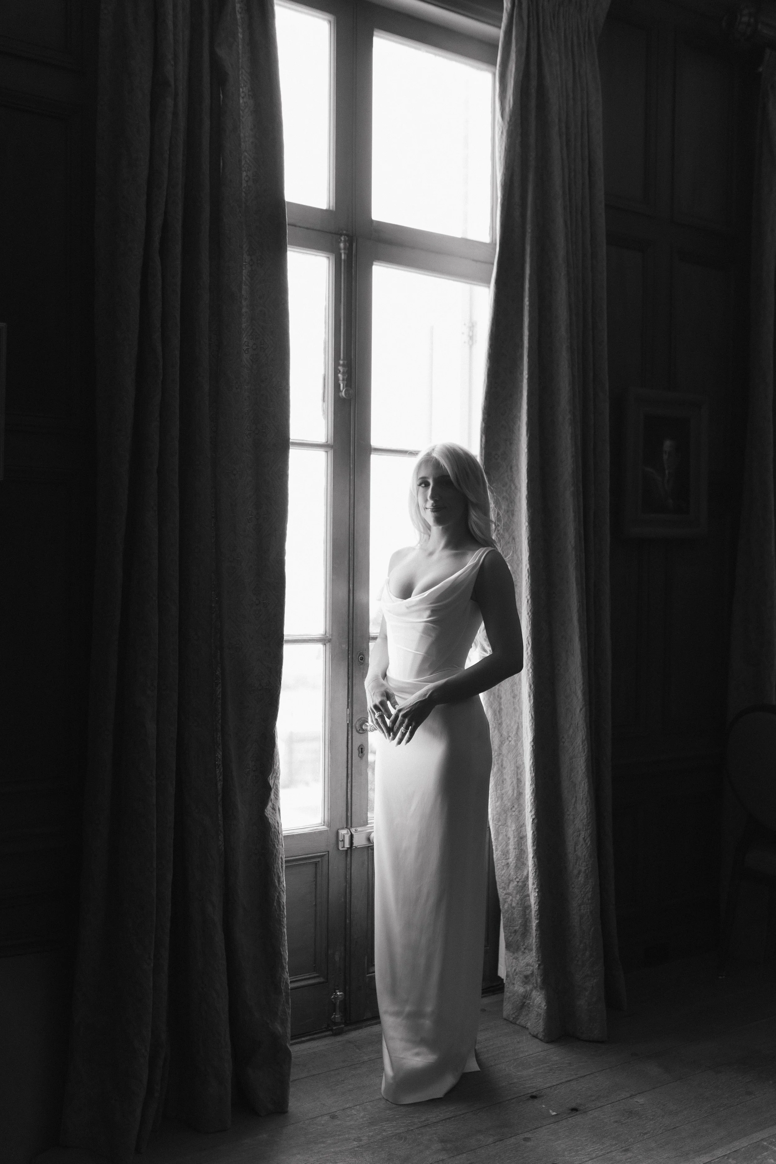 A woman in a long dress standing by a large window with curtains, in a room with wooden paneling, black and white photograph.