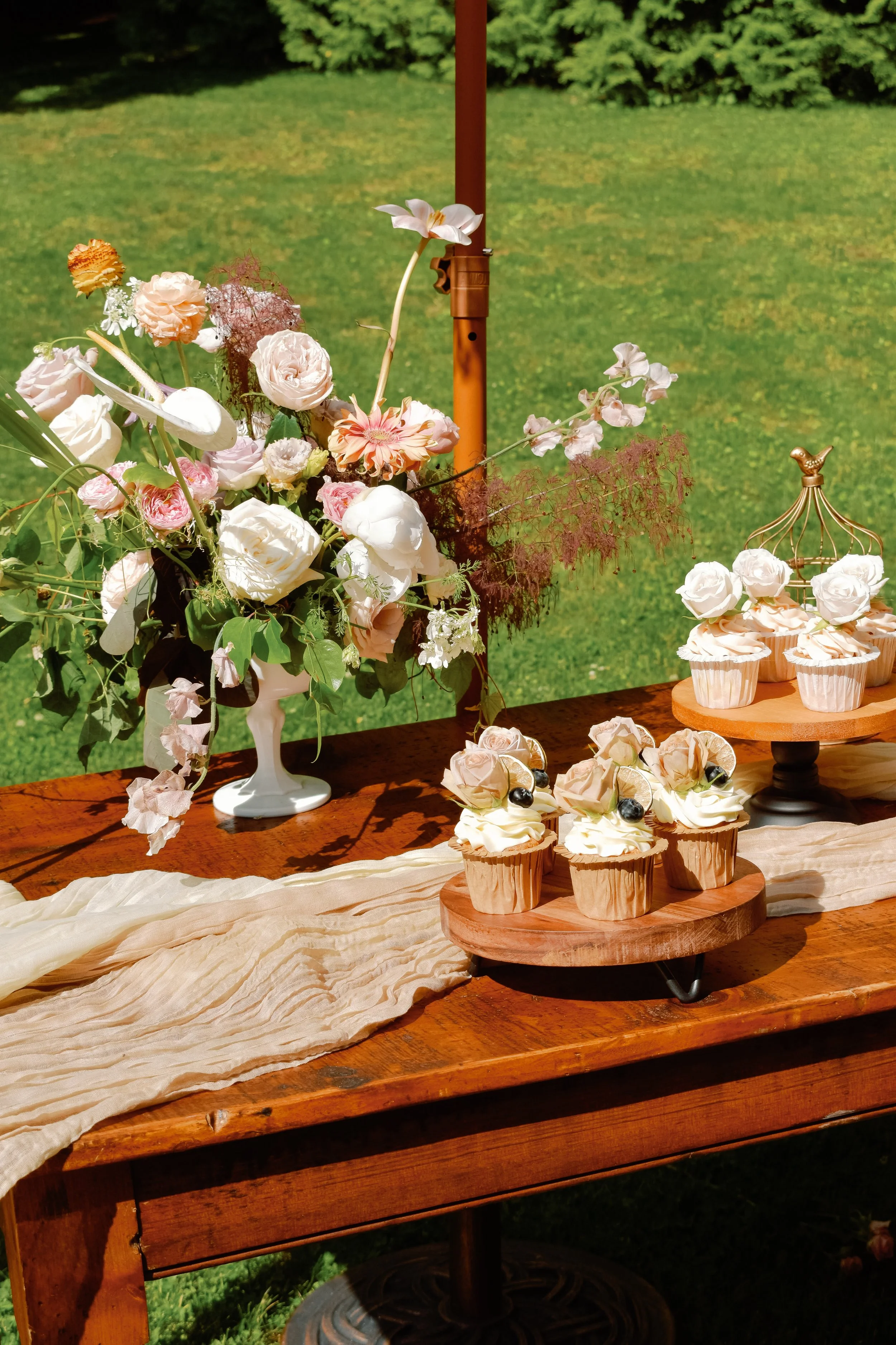 A table decorated with a beige cloth, a large floral arrangement in a white vase, and assorted cupcakes with frosting, flowers, and berries on wooden stands, set against a grassy outdoor backdrop.