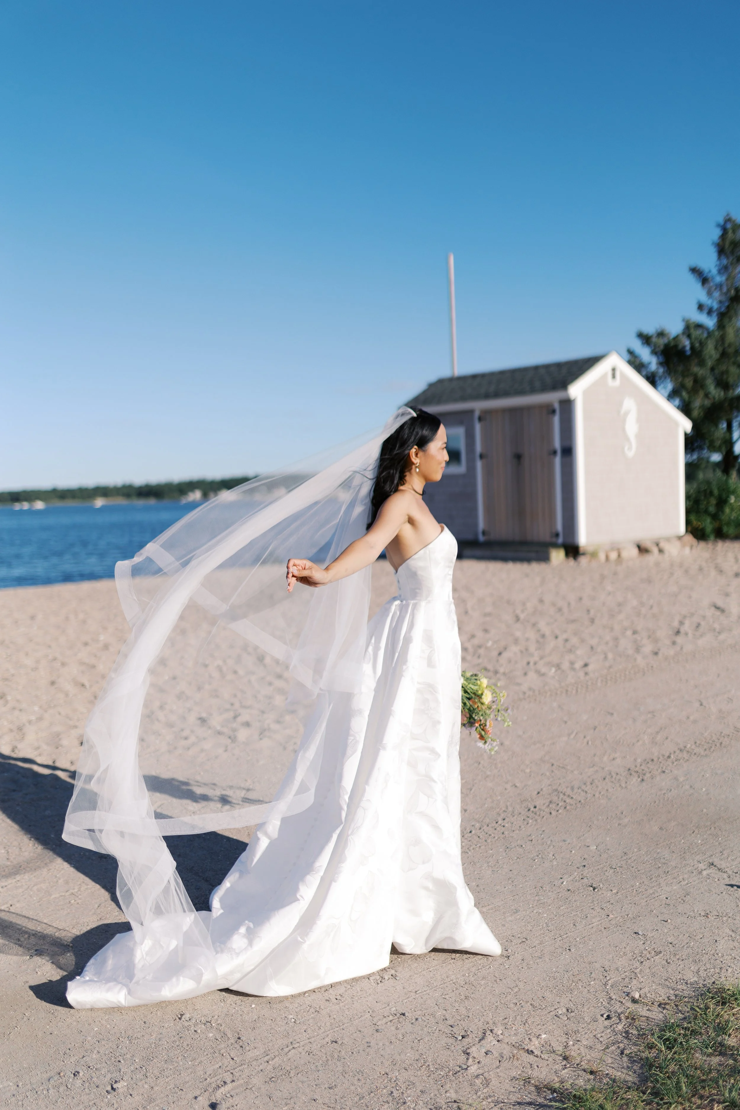 A bride in a white wedding dress with a veil standing on a sandy beach near a body of water with a small building in the background.
