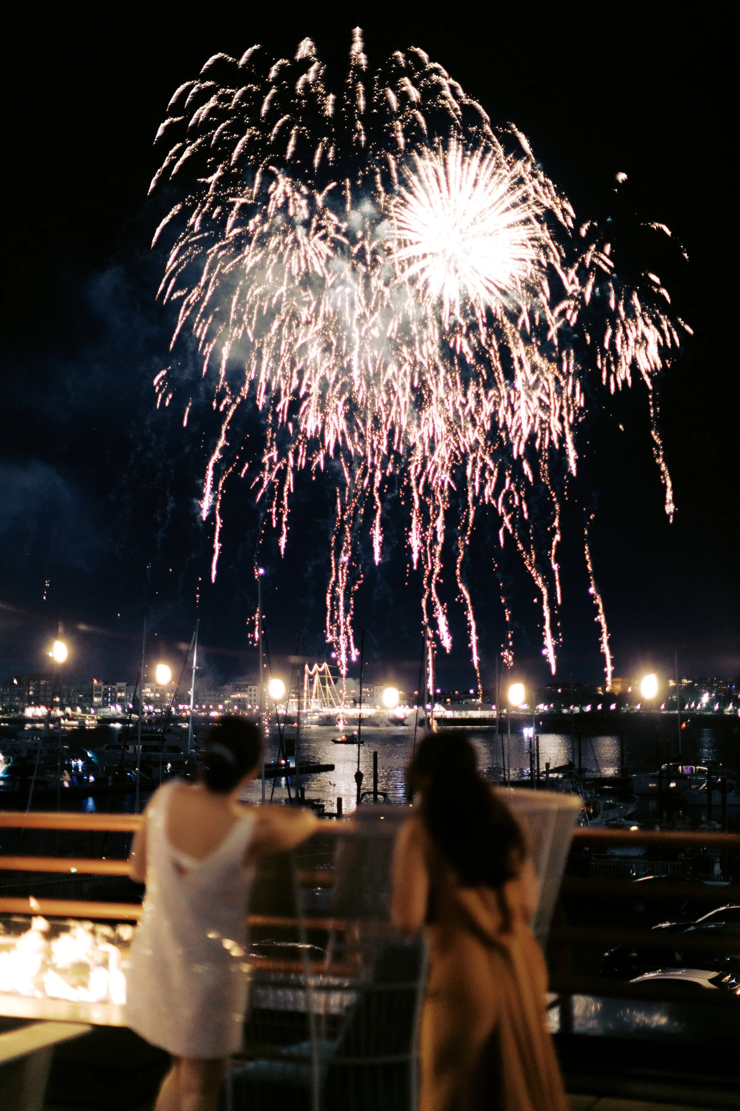 Two women watching fireworks over a harbor at night, with boats and cityscape in the background.