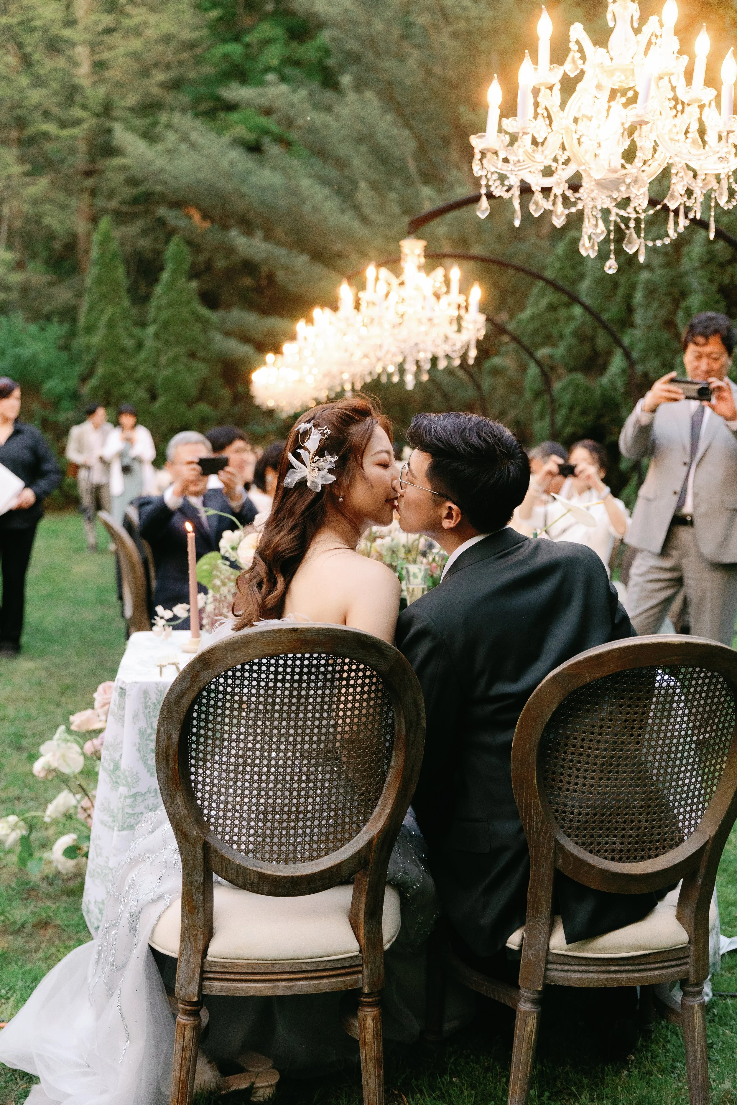A bride and groom sharing a kiss at their outdoor wedding reception, with chandeliers hanging above and guests taking photos in the background.