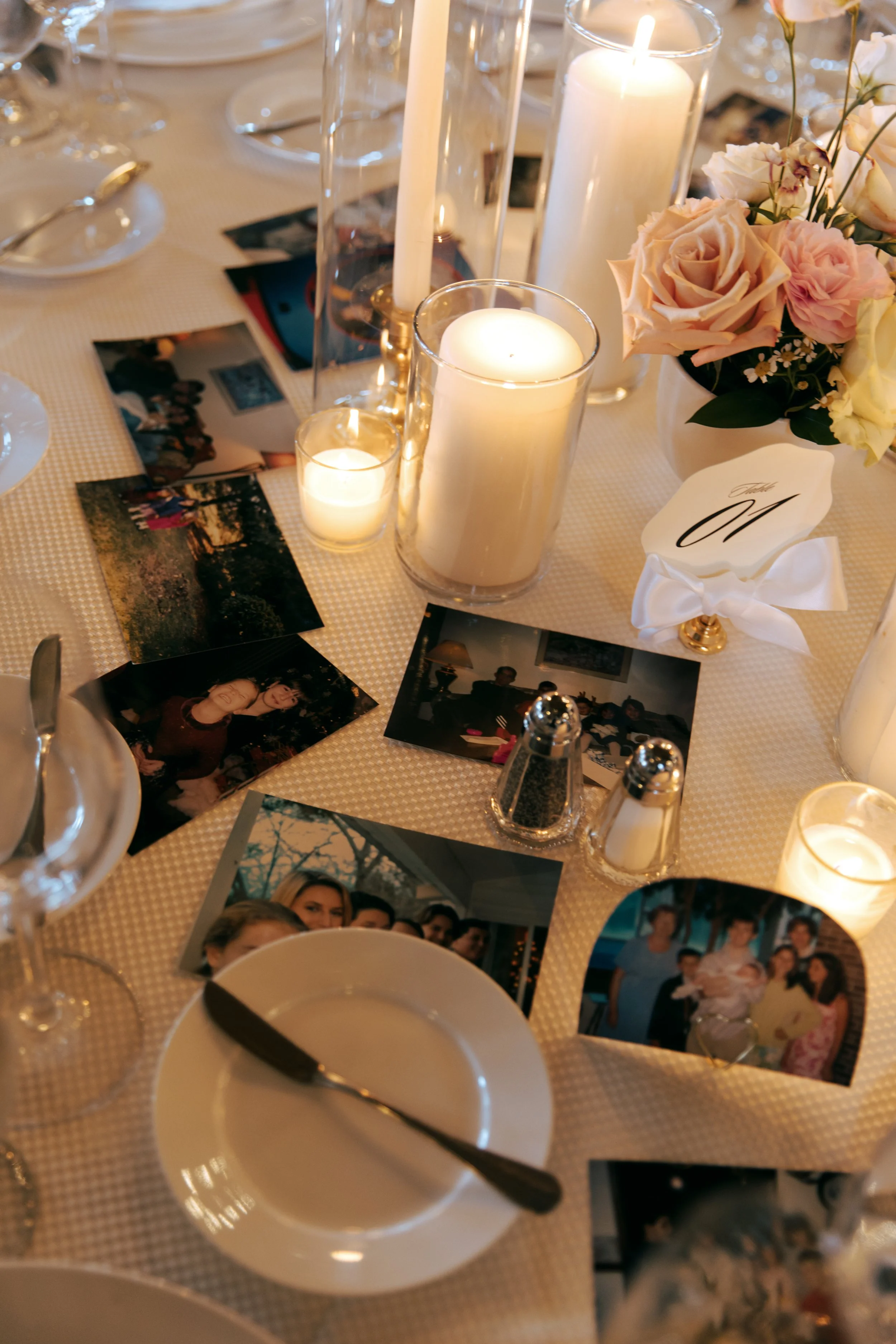 Table decorated with candles, a floral centerpiece, photographs, a white napkin with a gold 'M', and dinnerware at a celebration or gathering.