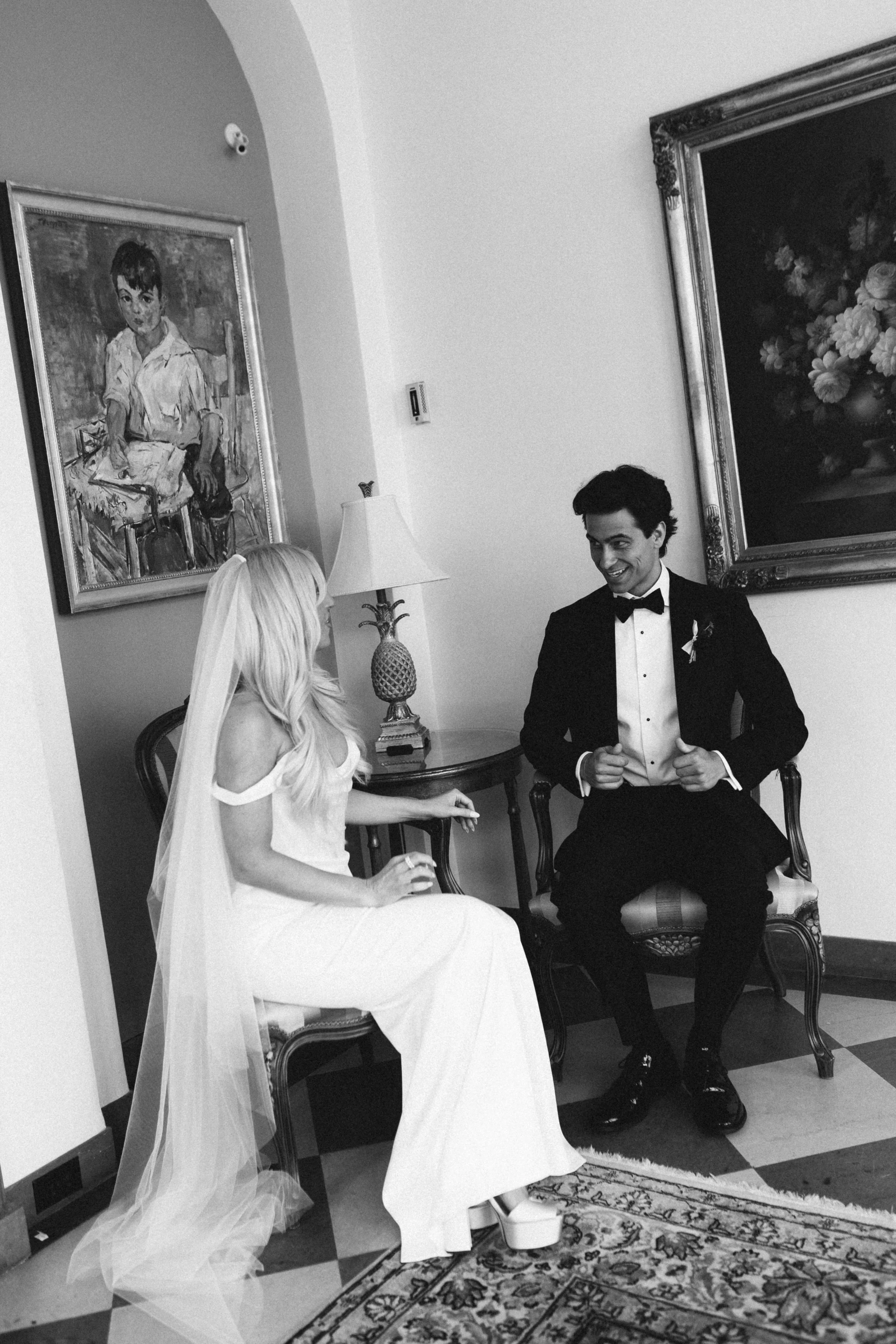 Bride and groom sitting indoors, the bride wearing a wedding dress with veil and the groom in a tuxedo, smiling at each other, with paintings and a table lamp in the background.