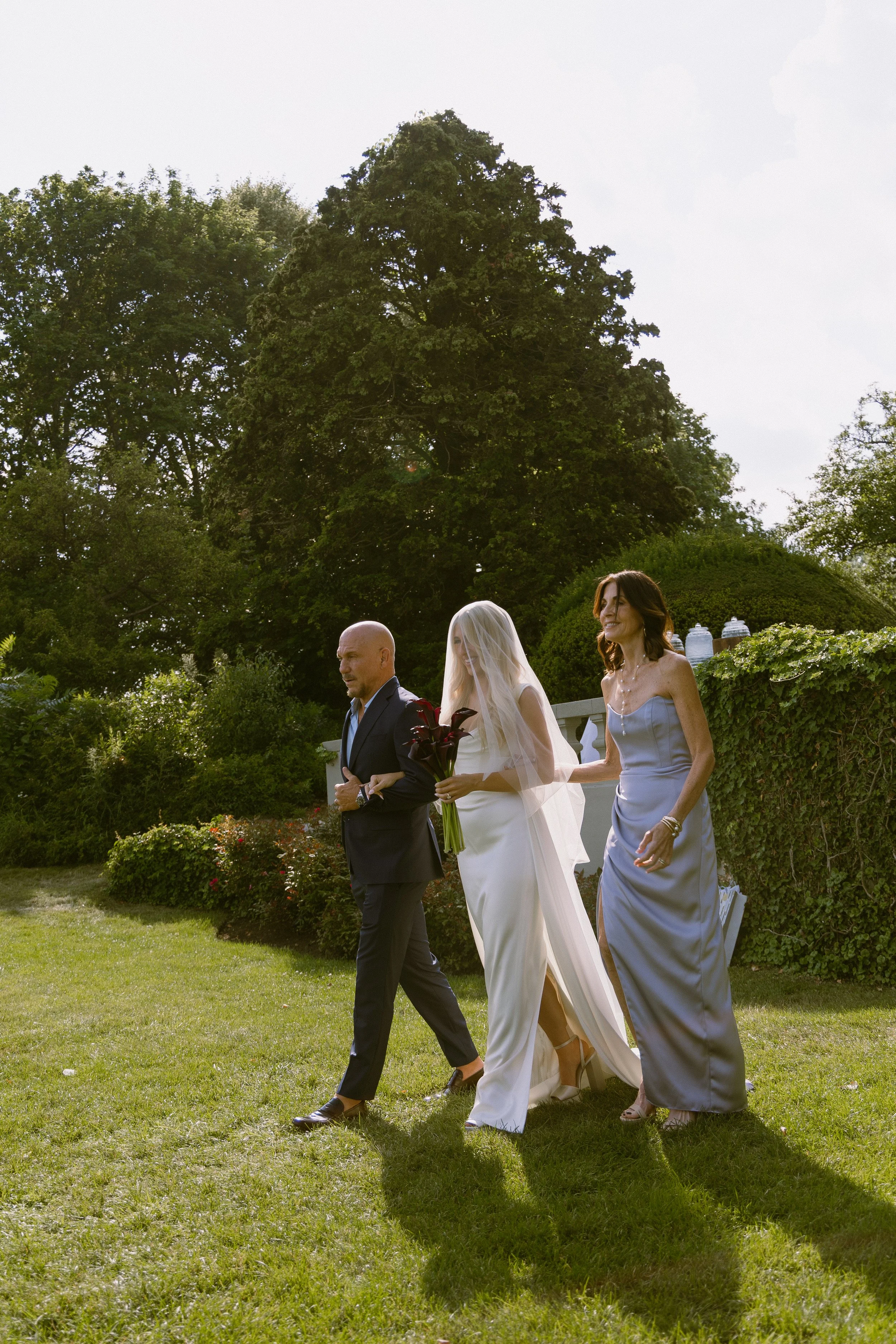 A bride in a white wedding dress and veil being escorted by a man in a dark suit, with a woman in a blue dress walking beside them in a garden setting during the day.