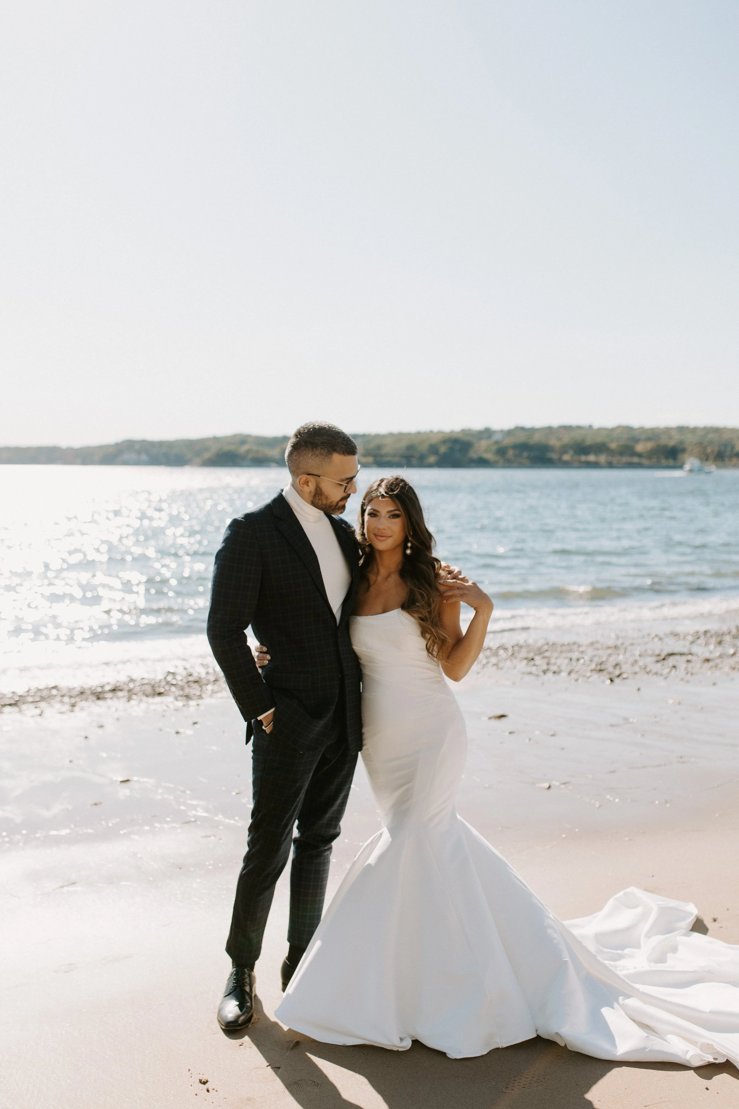 A bride and groom standing on the beach, sharing a moment near the water on their wedding day.