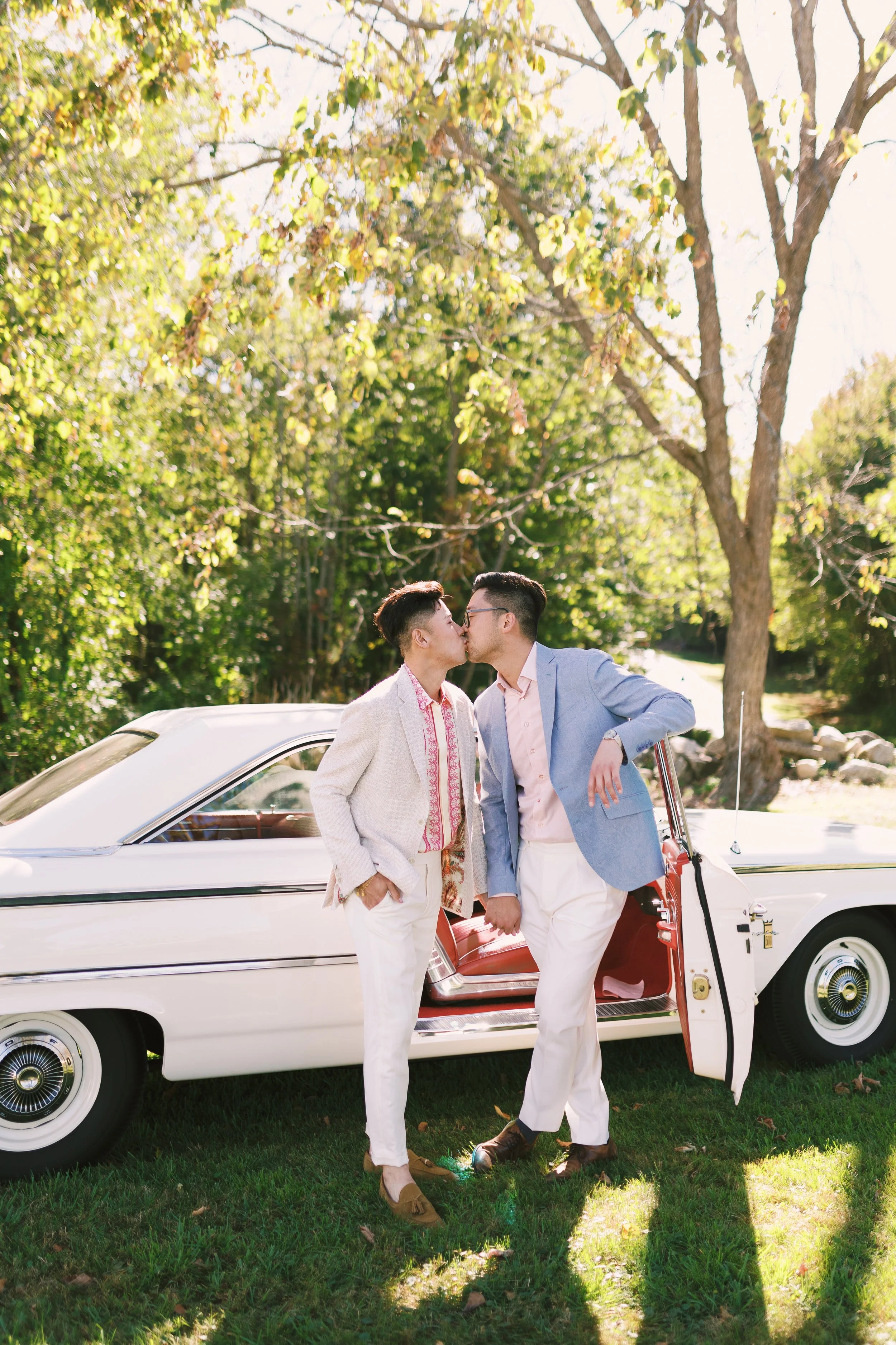 Two men wearing pastel-colored blazers and white pants, kissing, standing outside next to a vintage white car on a grassy area with trees and sunlight in the background.