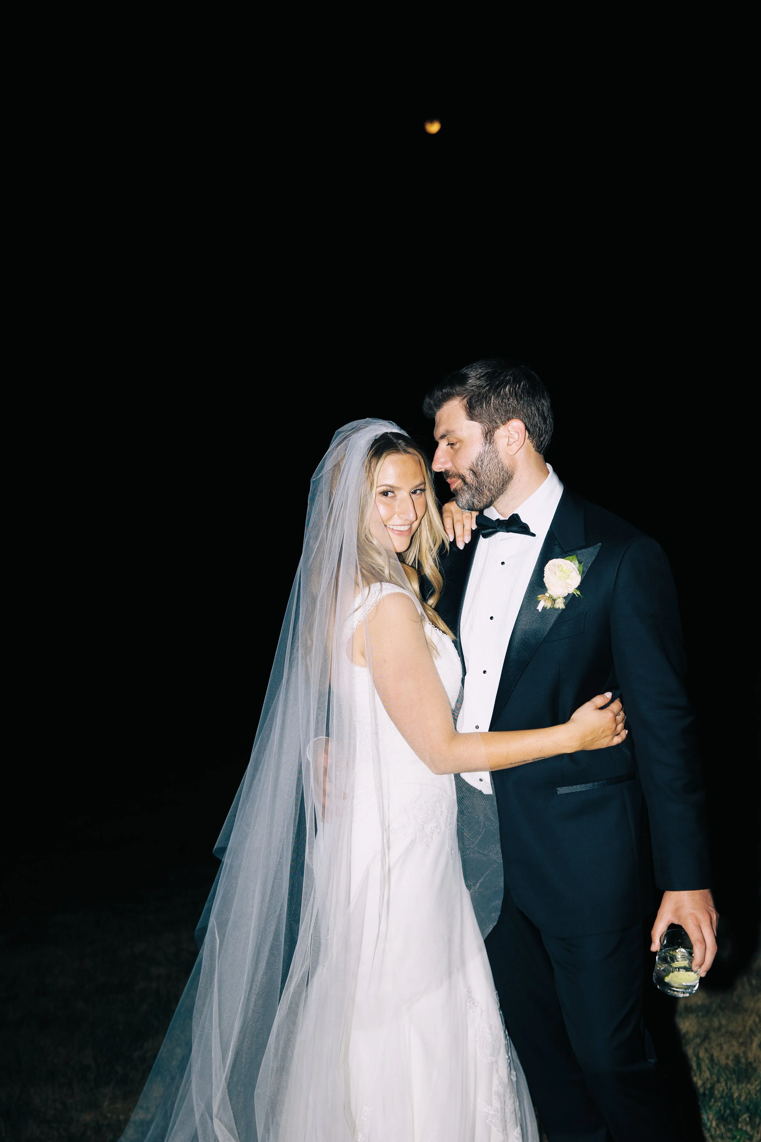 A bride and groom embrace during their wedding at night, with a dark sky and a visible moon in the background.
