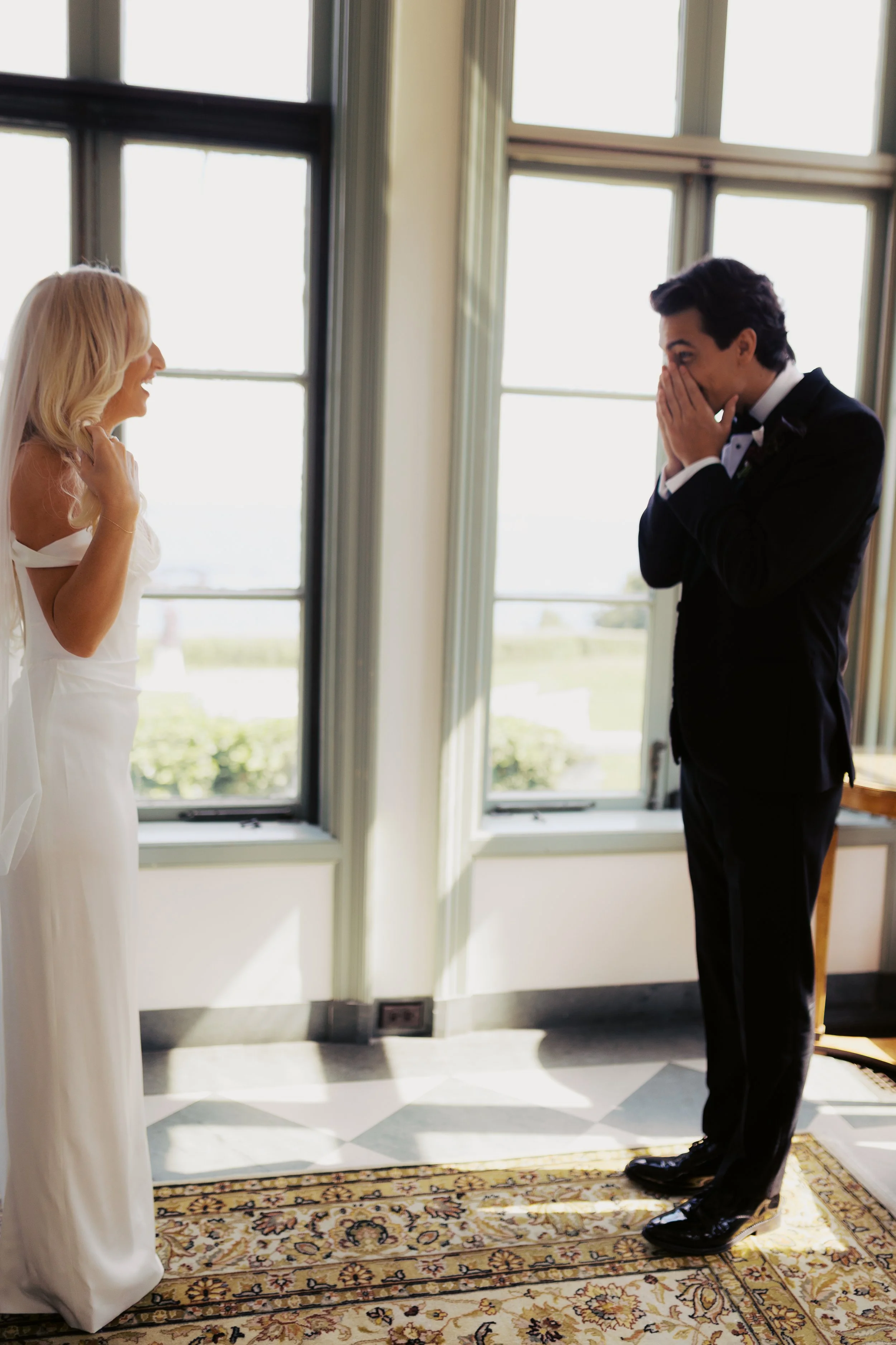 A bride and groom in formal attire standing in front of large windows, sharing a joyful moment with the bride laughing and the groom covering his face, possibly in an emotional reaction.
