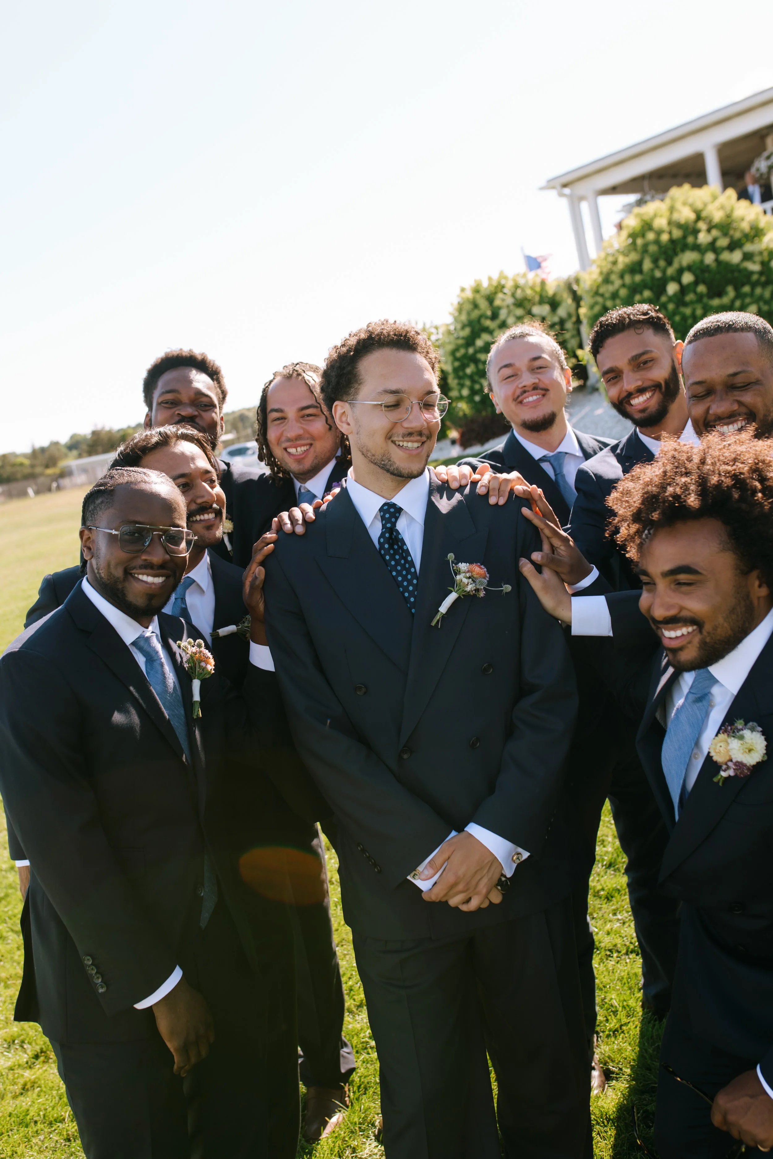 Group of men dressed in suits, smiling, and gathering outdoors on a sunny day, with a house and greenery in the background.