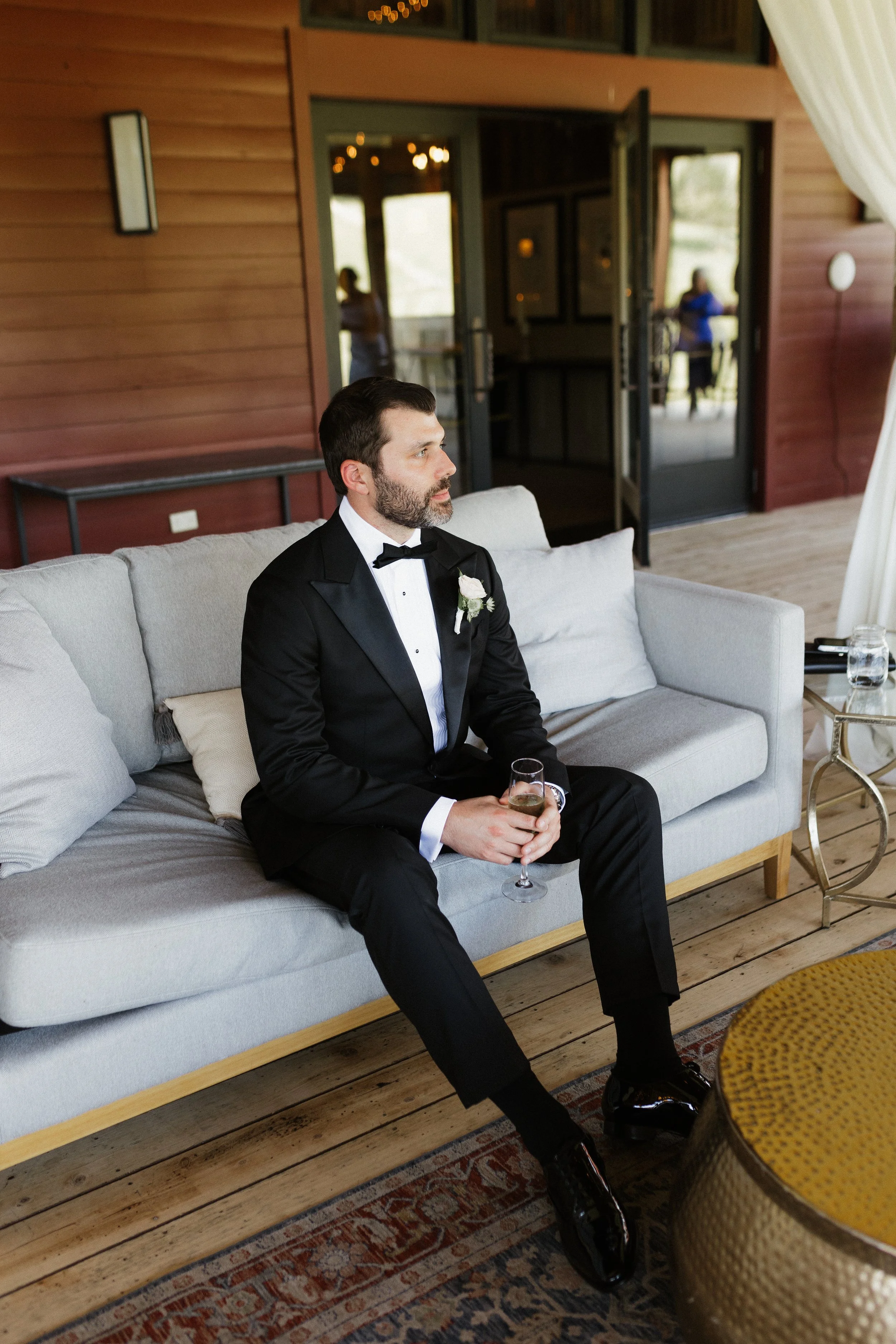 A man in a black tuxedo with a white shirt and black bow tie sitting on a light-colored sofa, holding a glass of champagne, with a wooden interior background.