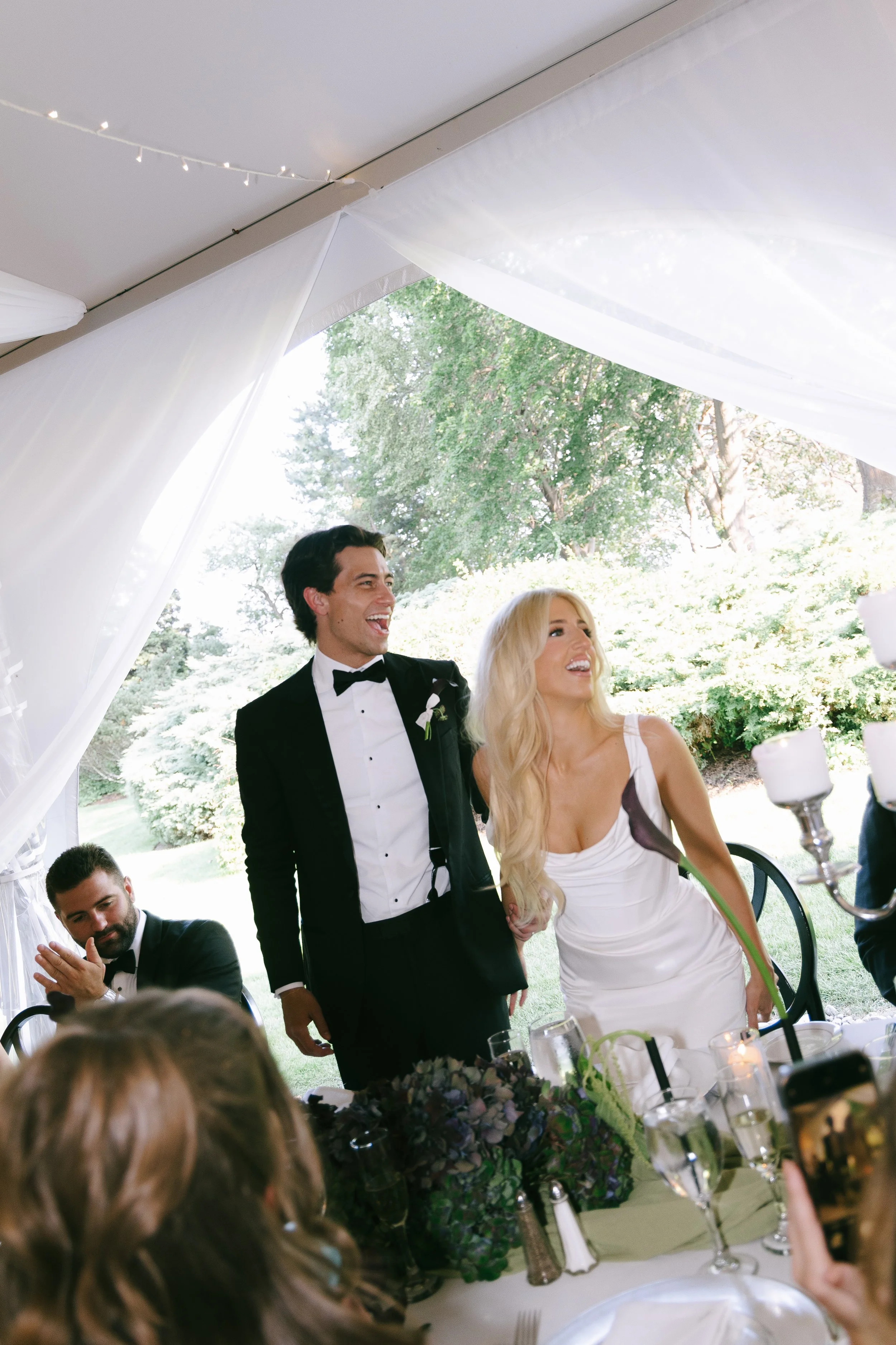 A newly married couple, a man in a tuxedo and a woman in a white wedding dress, standing together and smiling at a wedding reception outdoors under a white tent.