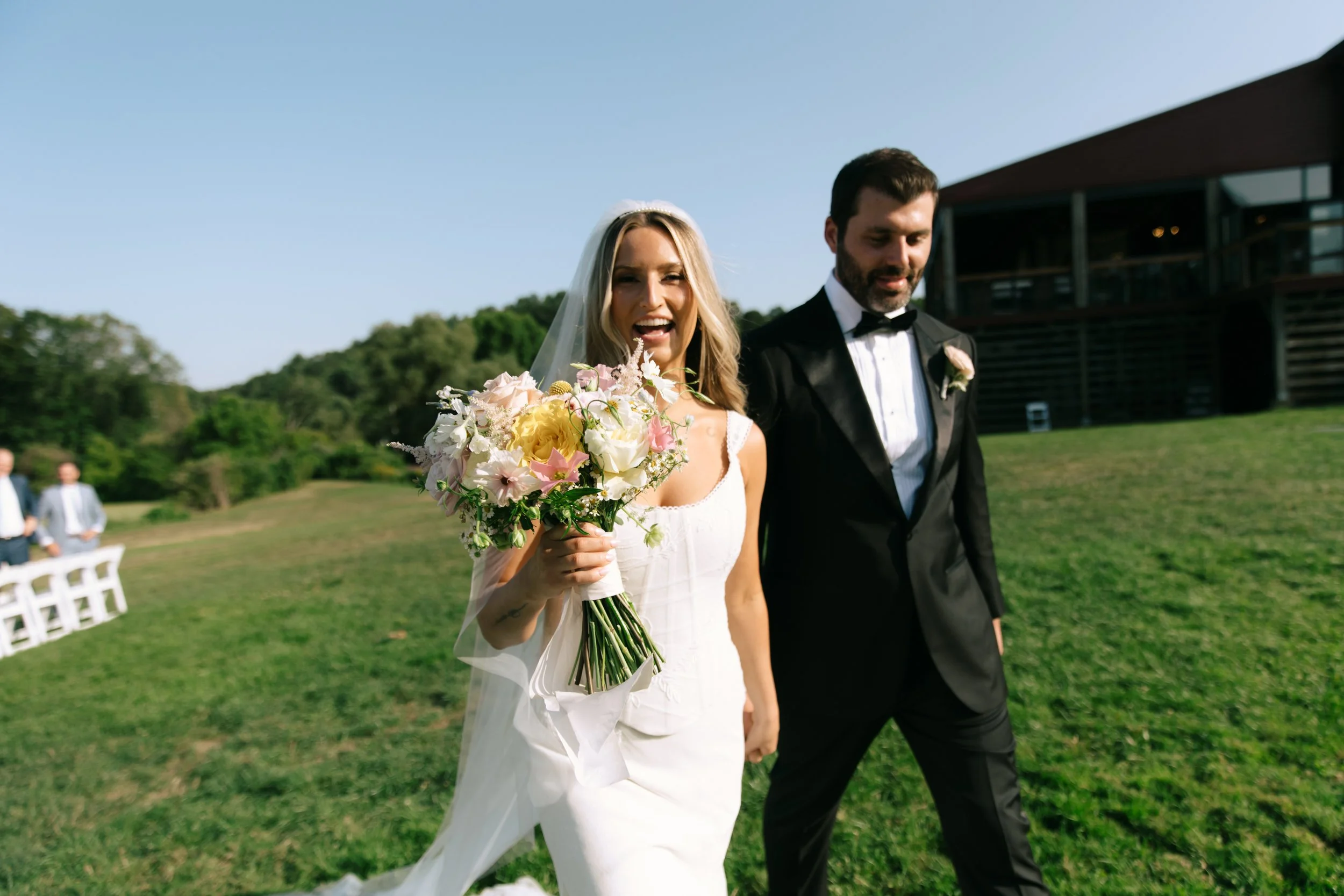 Bride and groom walking outside during wedding ceremony, bride holding a bouquet of flowers, bride in white dress with veil, groom in black tuxedo with bow tie, green landscape, modern wooden building in background.