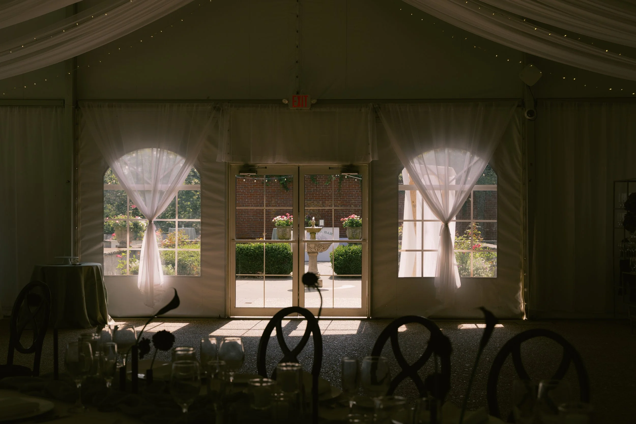 Interior view of a decorated event tent with sheer curtains and chairs, overlooking a patio with potted flowers and a brick wall outside.