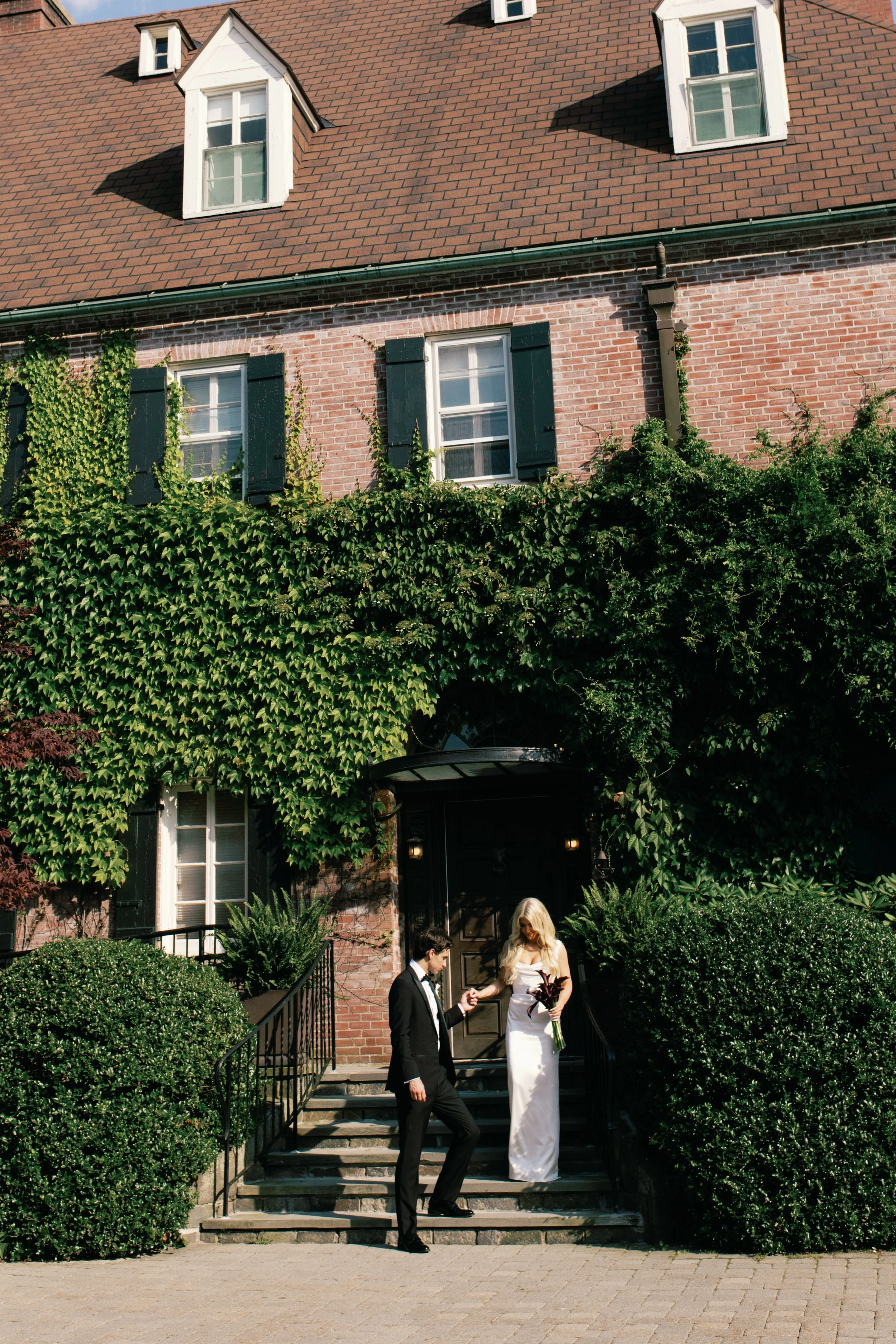 A bride and groom standing on steps outside a brick house with green shutters, surrounded by lush green bushes and ivy.