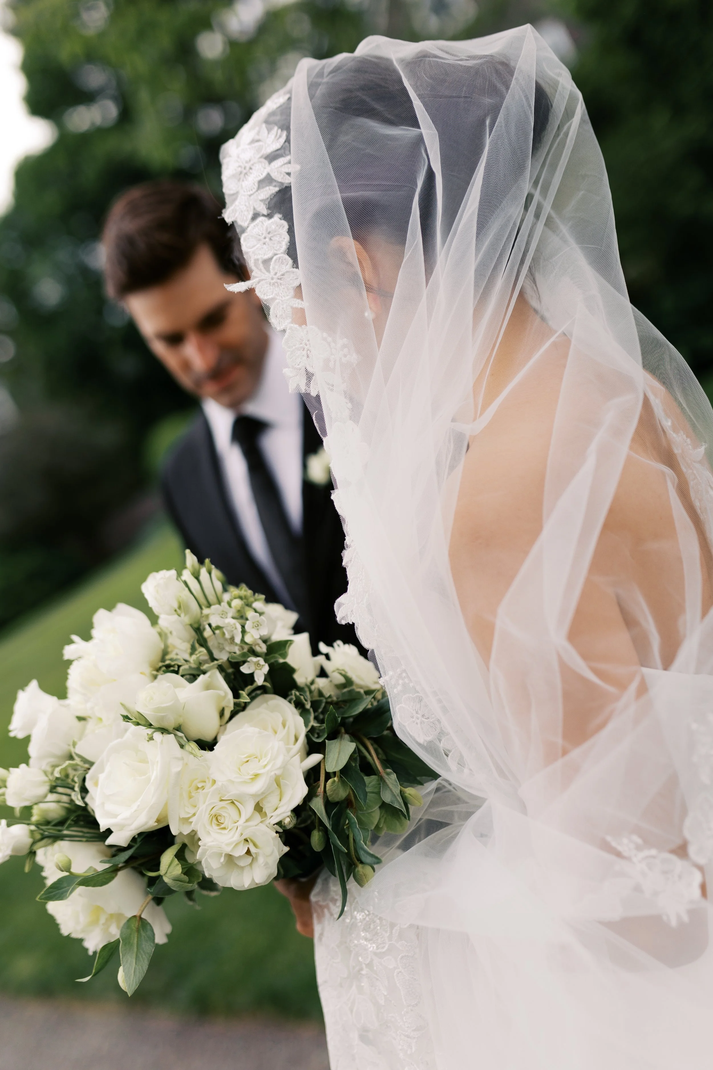 Bride in a white veil holding a bouquet of white roses and greenery, standing outdoors with a groom in a black suit and tie in the background.