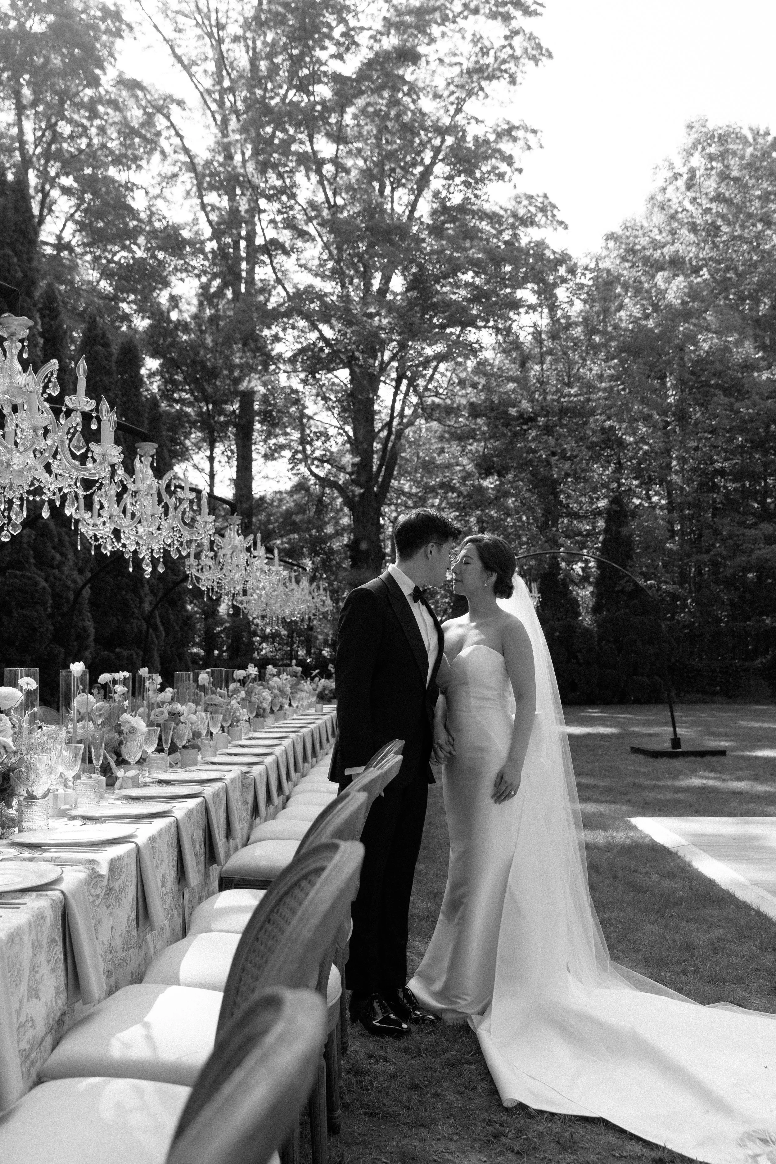 A bride and groom stand close together outdoors on their wedding day, with the bride wearing a strapless gown and veil, and the groom in a tuxedo, amid trees and decorated tables.