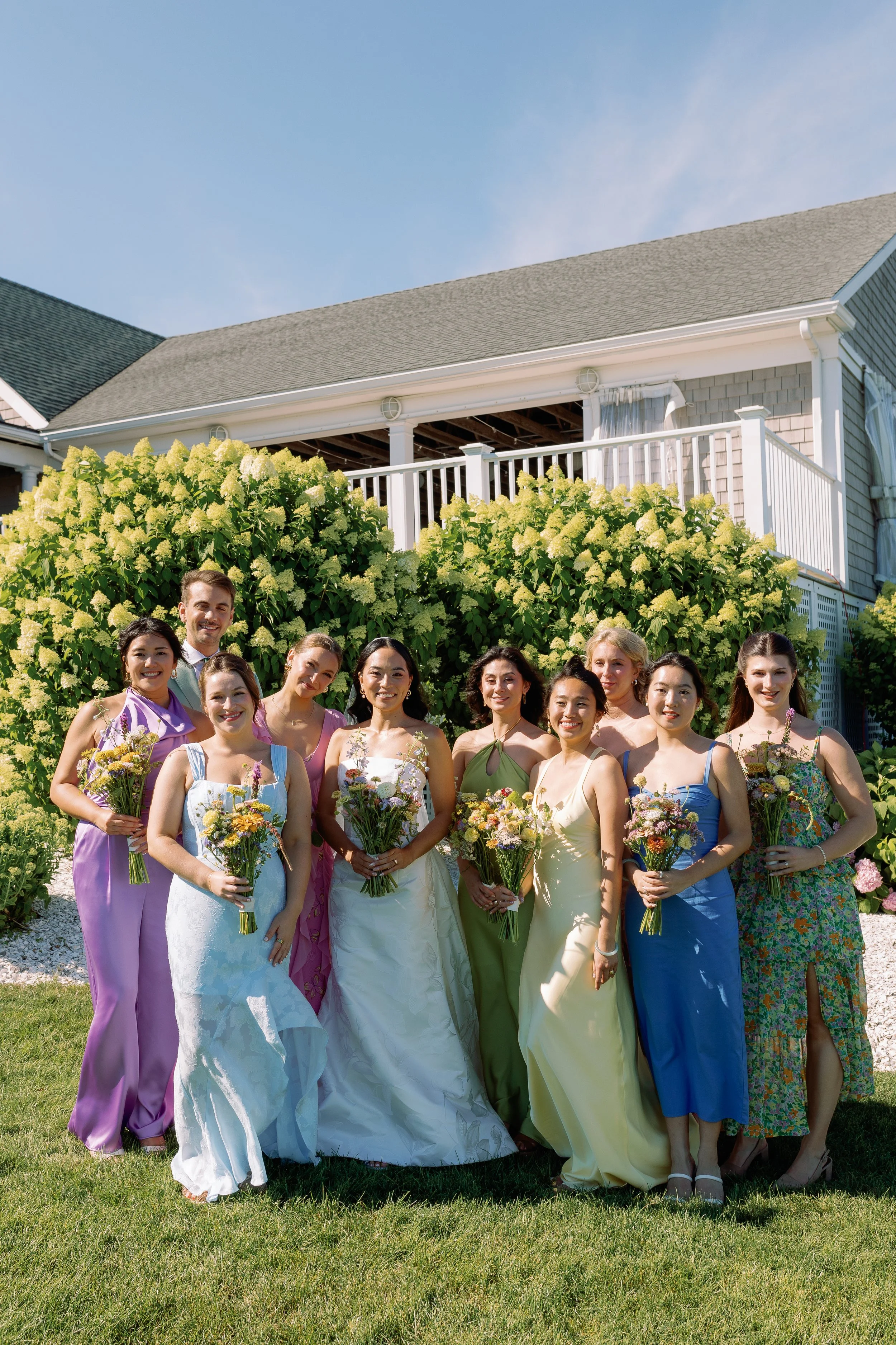 A group of women in colorful dresses holding bouquets of flowers, standing outdoors on grass, with a large bush and house in the background.
