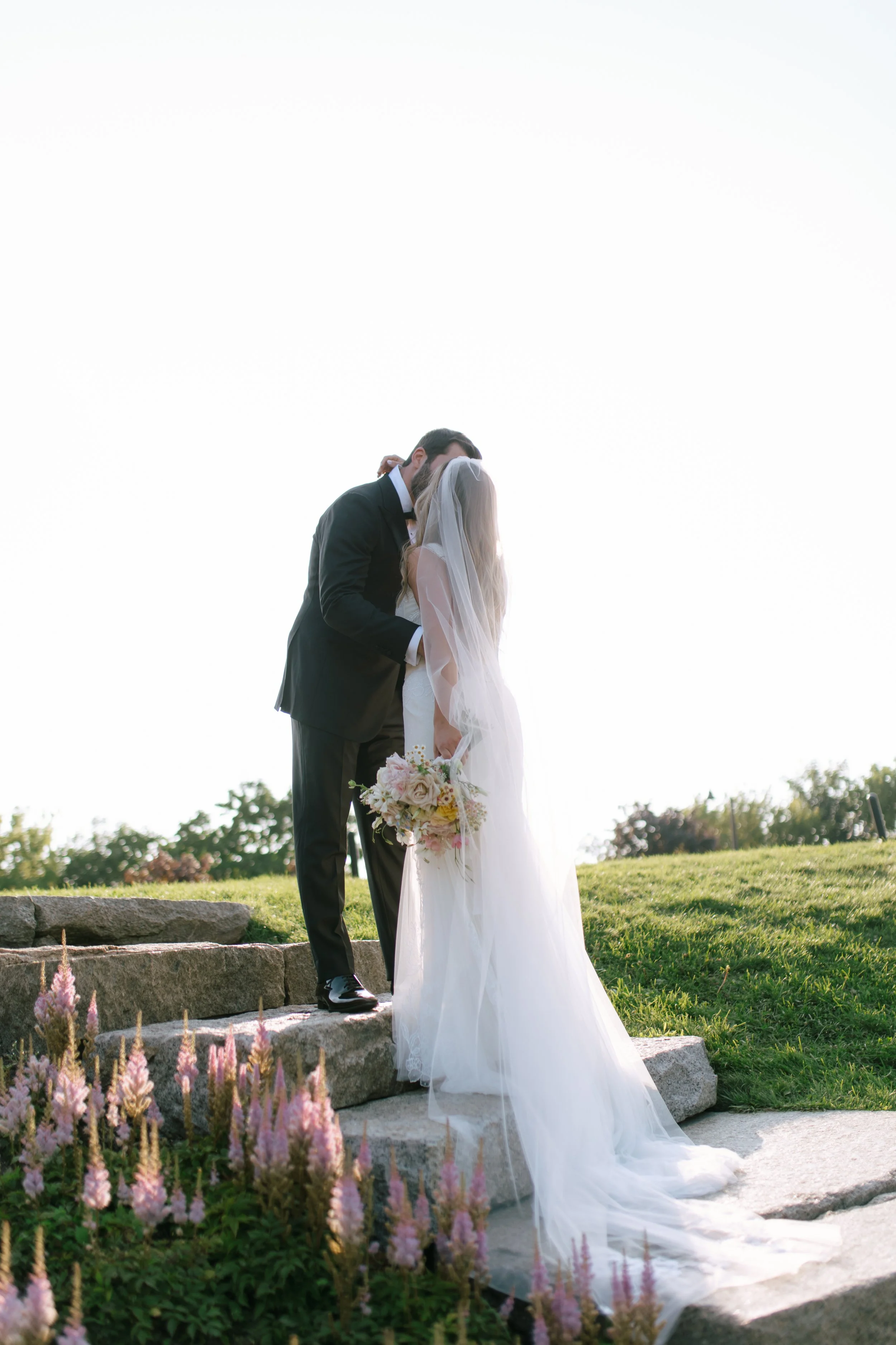 A bride and groom share a kiss outdoors on stone steps during sunset, with the bride holding a bouquet and wearing a long white gown and veil.