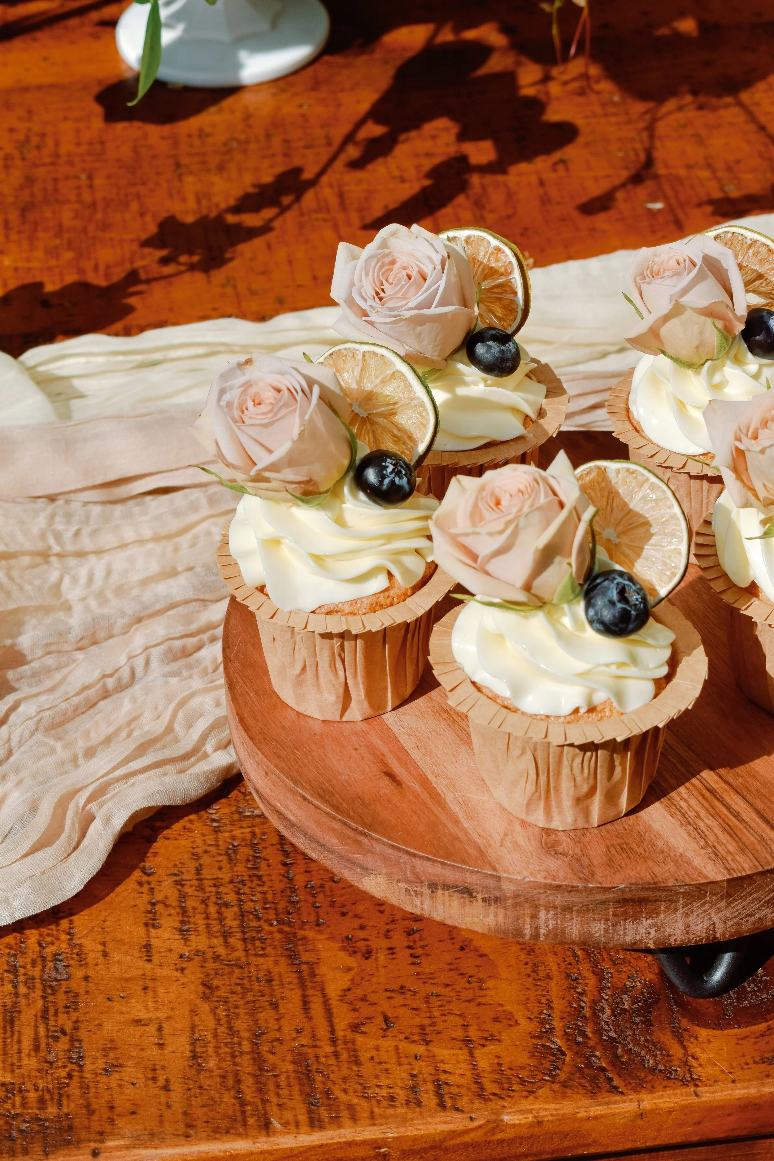 Cupcakes decorated with pink roses, dried lemon slices, and blueberries on a wooden platter.