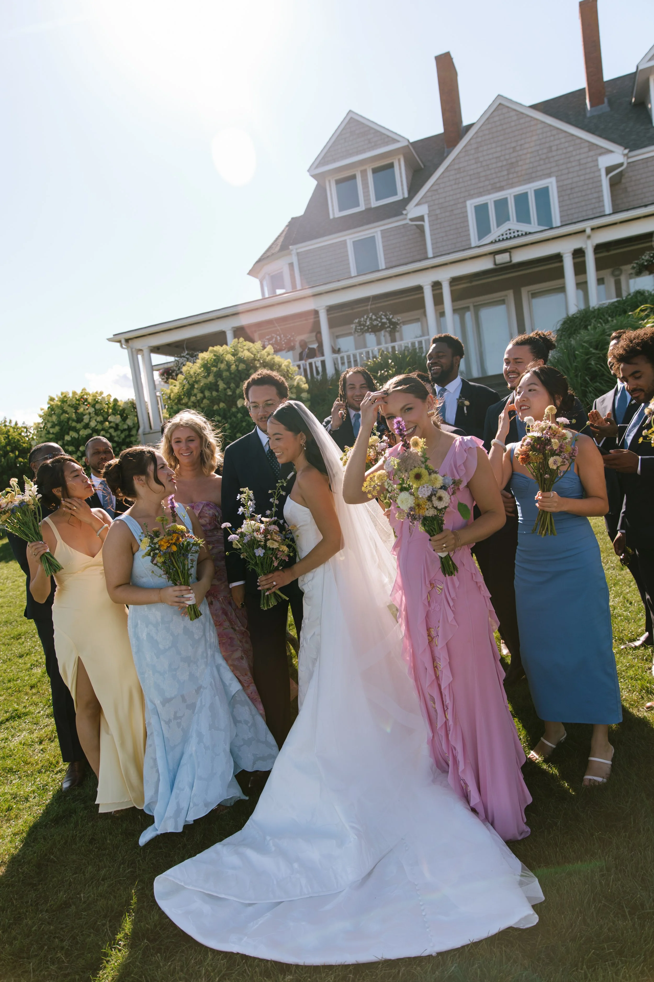 A wedding party gathers outside on a sunny day, with a large house in the background. The bride and groom are surrounded by bridesmaids and groomsmen holding bouquets of flowers, smiling and celebrating.