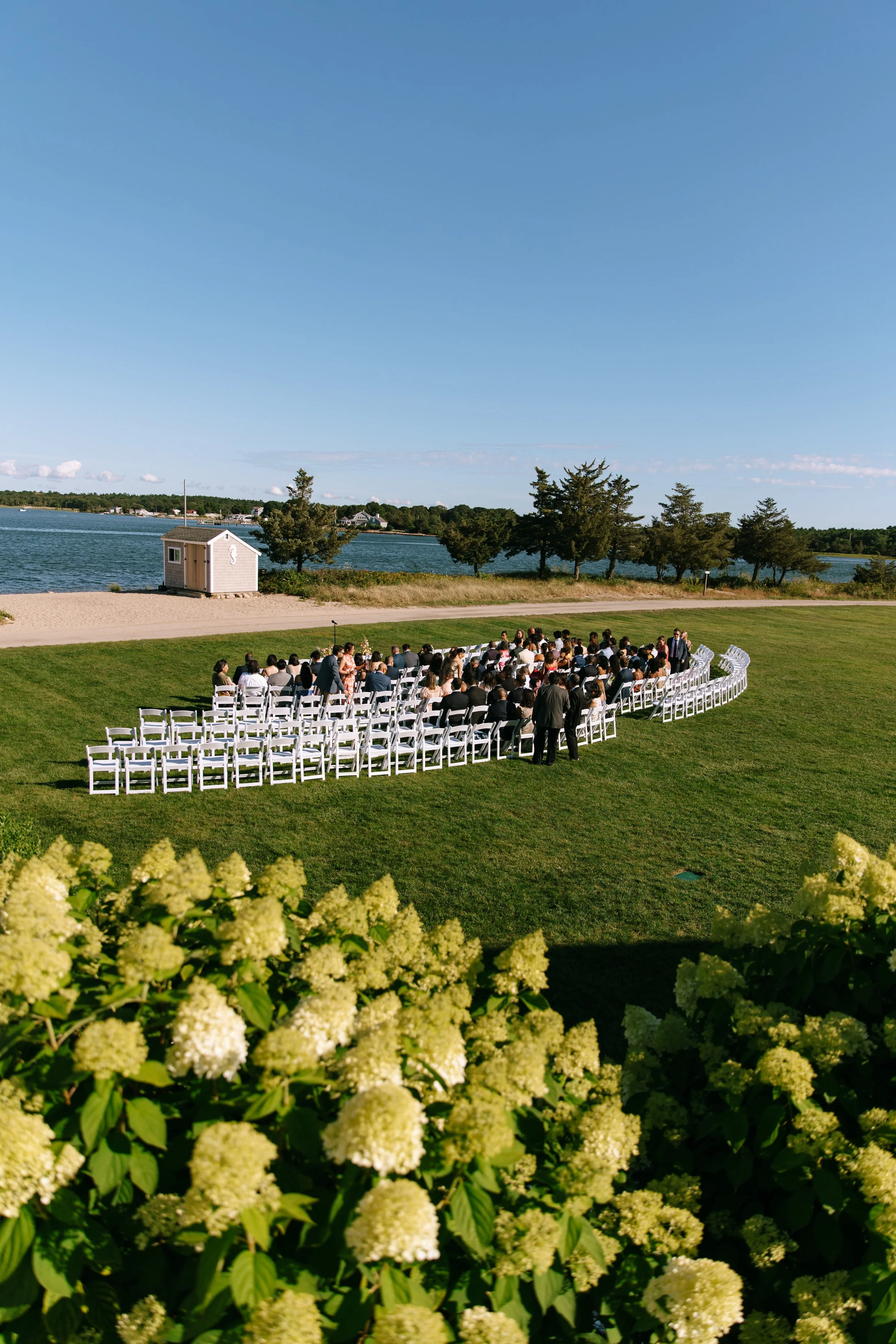 An outdoor wedding ceremony setup near a body of water with rows of white chairs on a grassy area, and a group of people gathered in the middle, under a clear blue sky with trees and a small shed nearby.