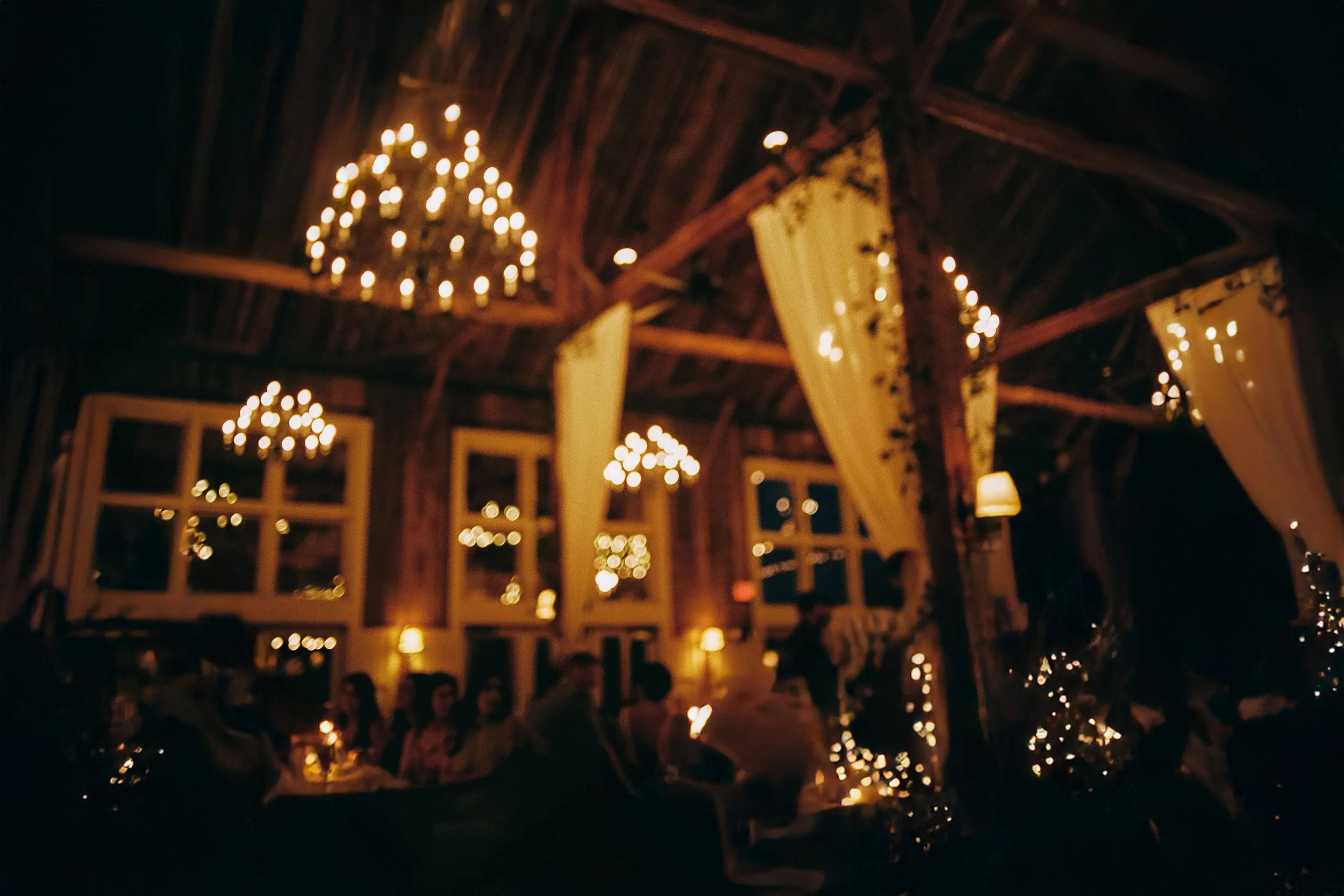 A dimly lit indoor event space with wooden ceilings, chandeliers with multiple small lights, and yellow curtains. Several people are seated at tables, and the room is decorated with string lights and lamps.