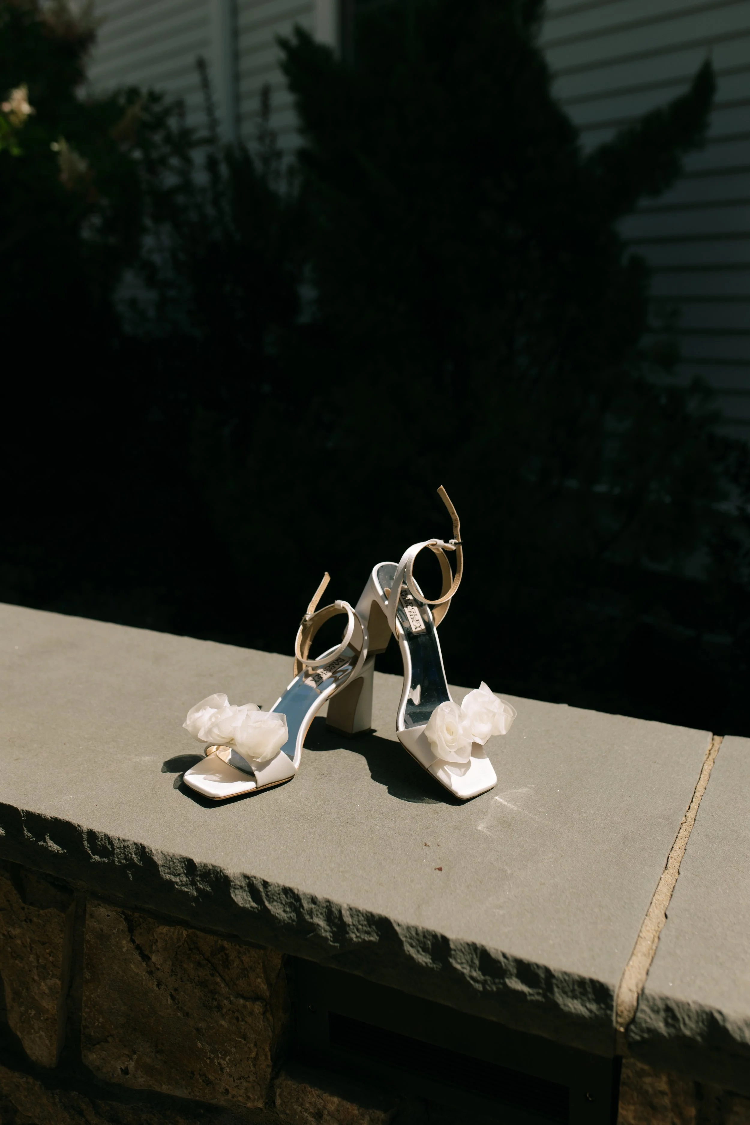 A pair of white high-heeled sandals with decorative white flowers on the front, placed on a stone surface outdoors, with a dark background.