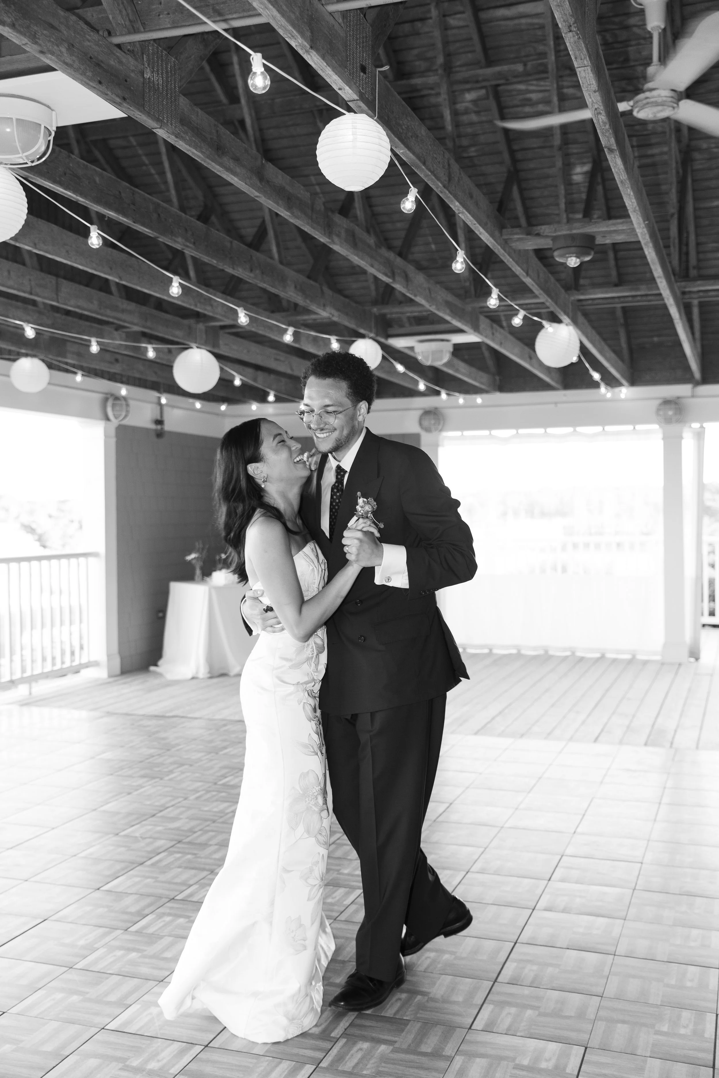 A couple dancing, dressed in wedding attire, in a decorated hall with paper lanterns and string lights hanging from the ceiling, during a wedding reception.