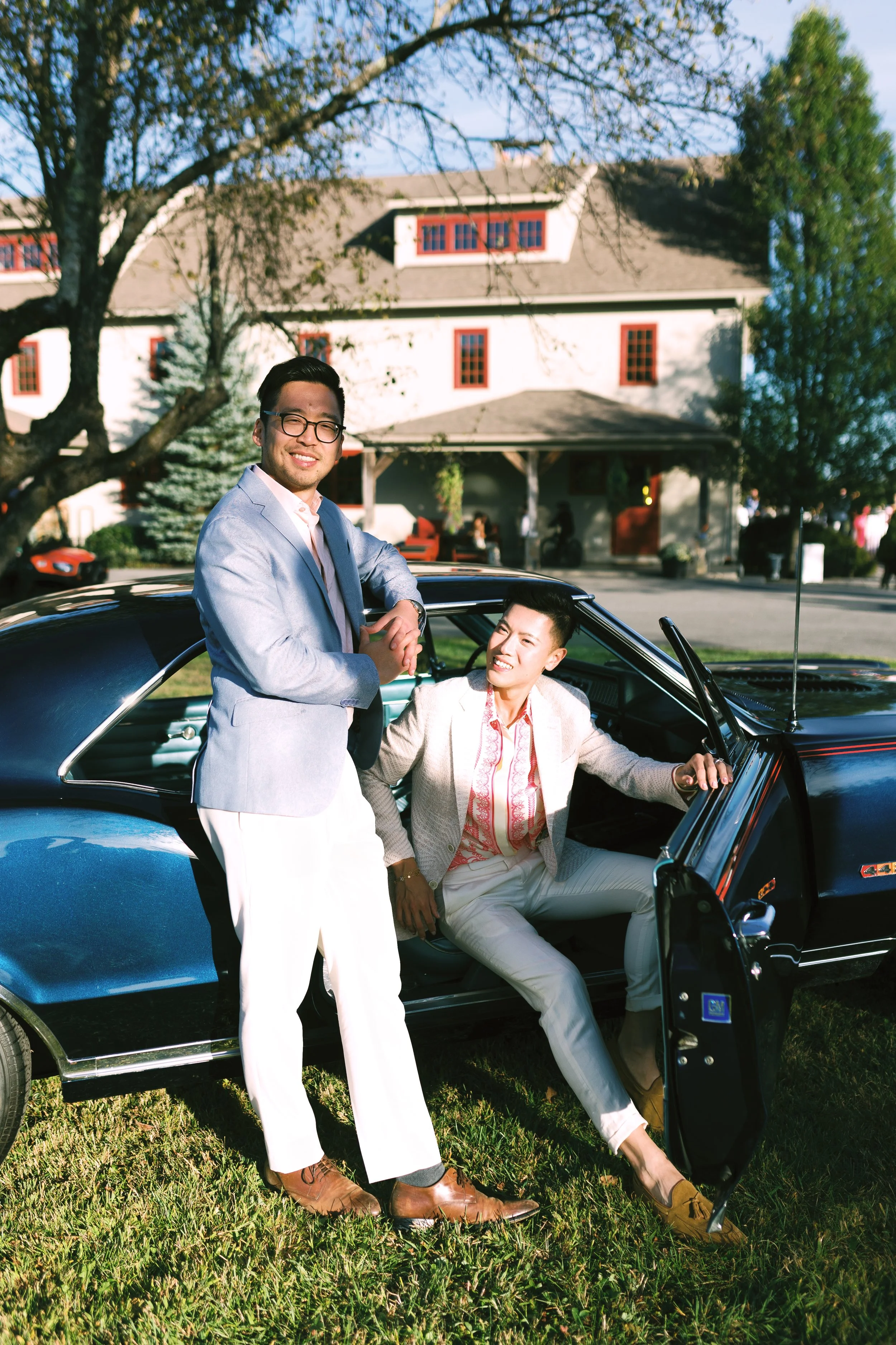 Two men in stylish clothing, one standing and one sitting in a vintage black car, outdoors with a large house and trees in the background.